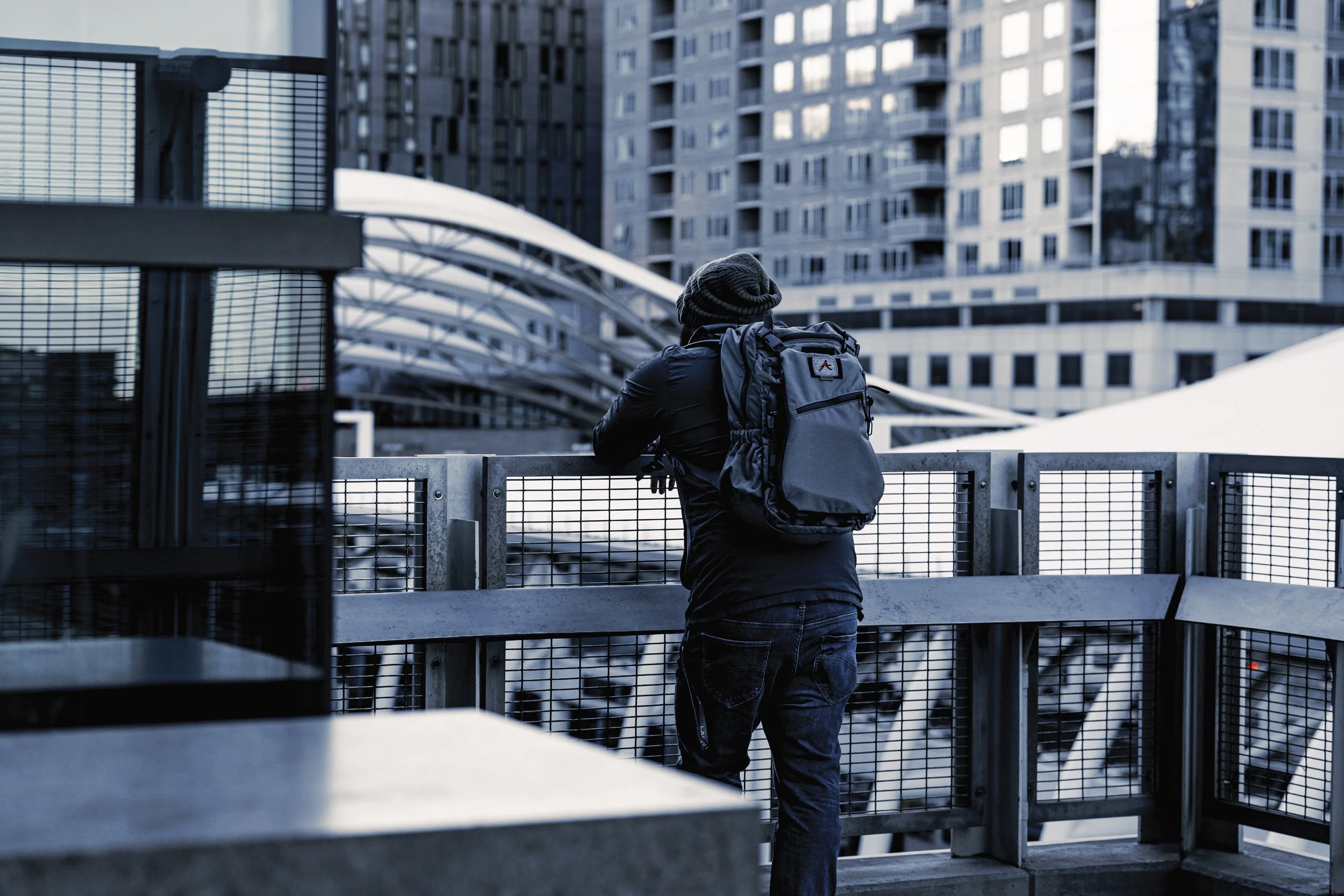 Person with a backpack looking over a city balcony, with modern high-rise buildings in the background.