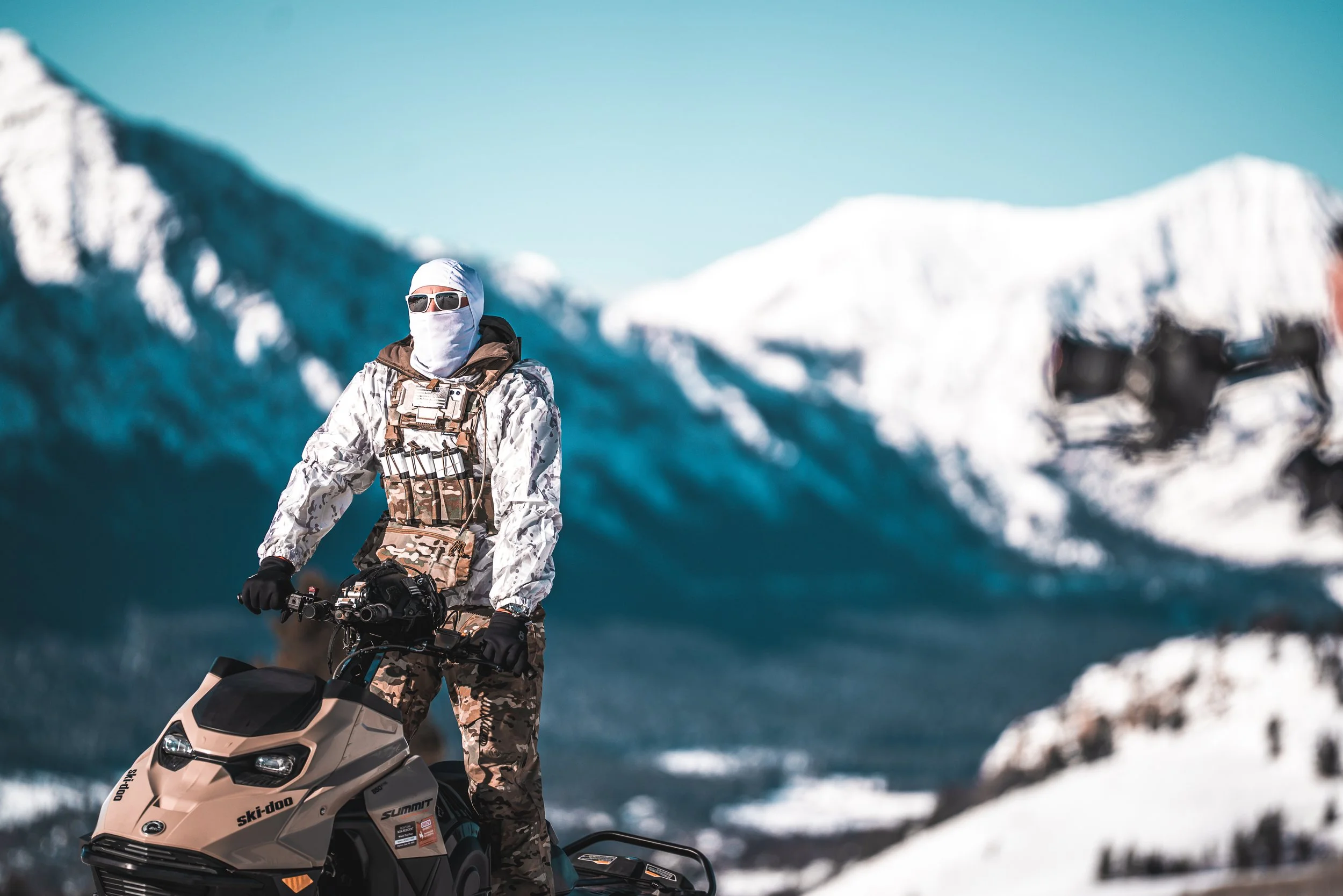 Person wearing camouflage outfit riding a snowmobile in snowy mountain landscape.