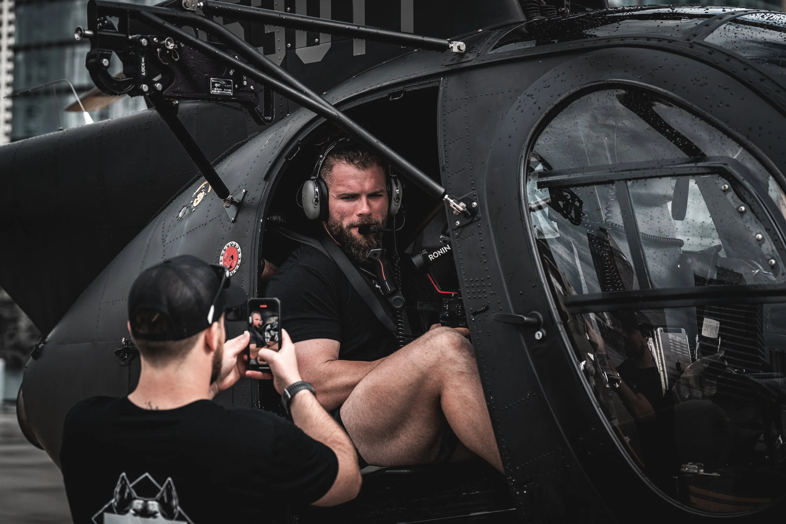 A man with a beard and muscular build sitting inside a black helicopter, wearing a headset, while another person takes a photo or video of him with a smartphone.