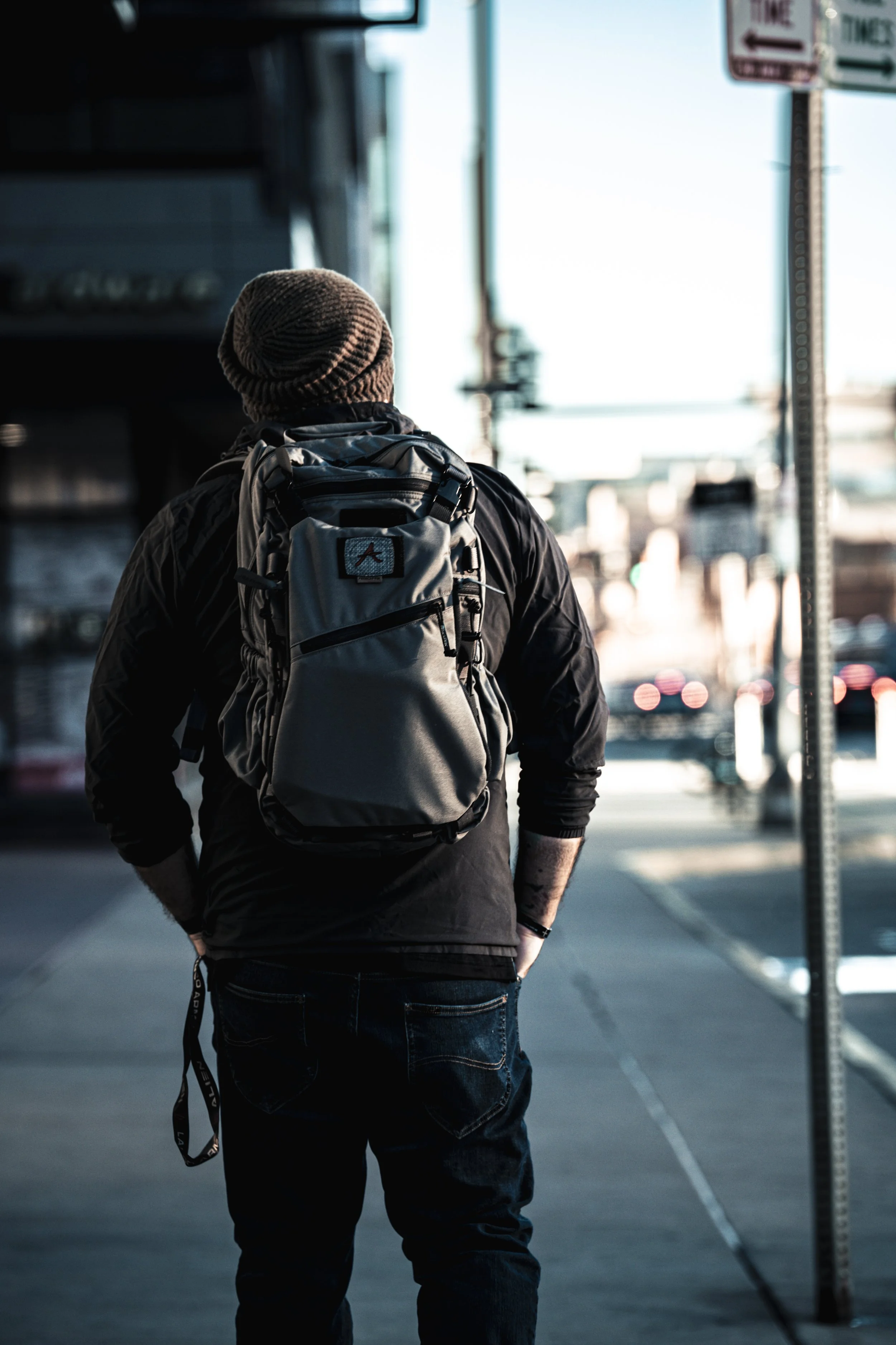 A person wearing a beanie and black jacket with a large gray backpack standing on a city sidewalk, facing away, with blurred street scene in the background.