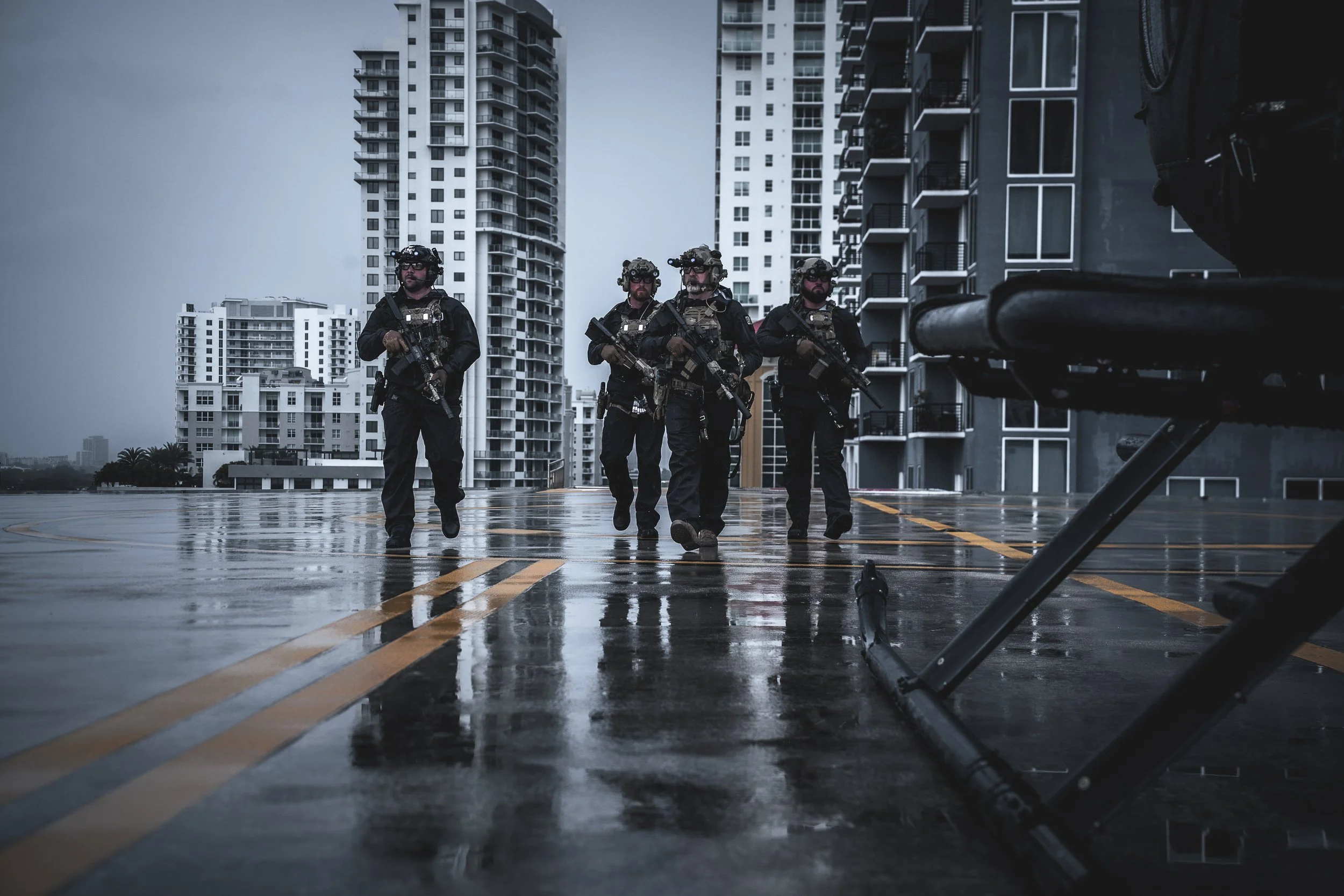 Four heavily armed tactical police officers walking on a wet city street during overcast weather with tall apartment buildings in the background.