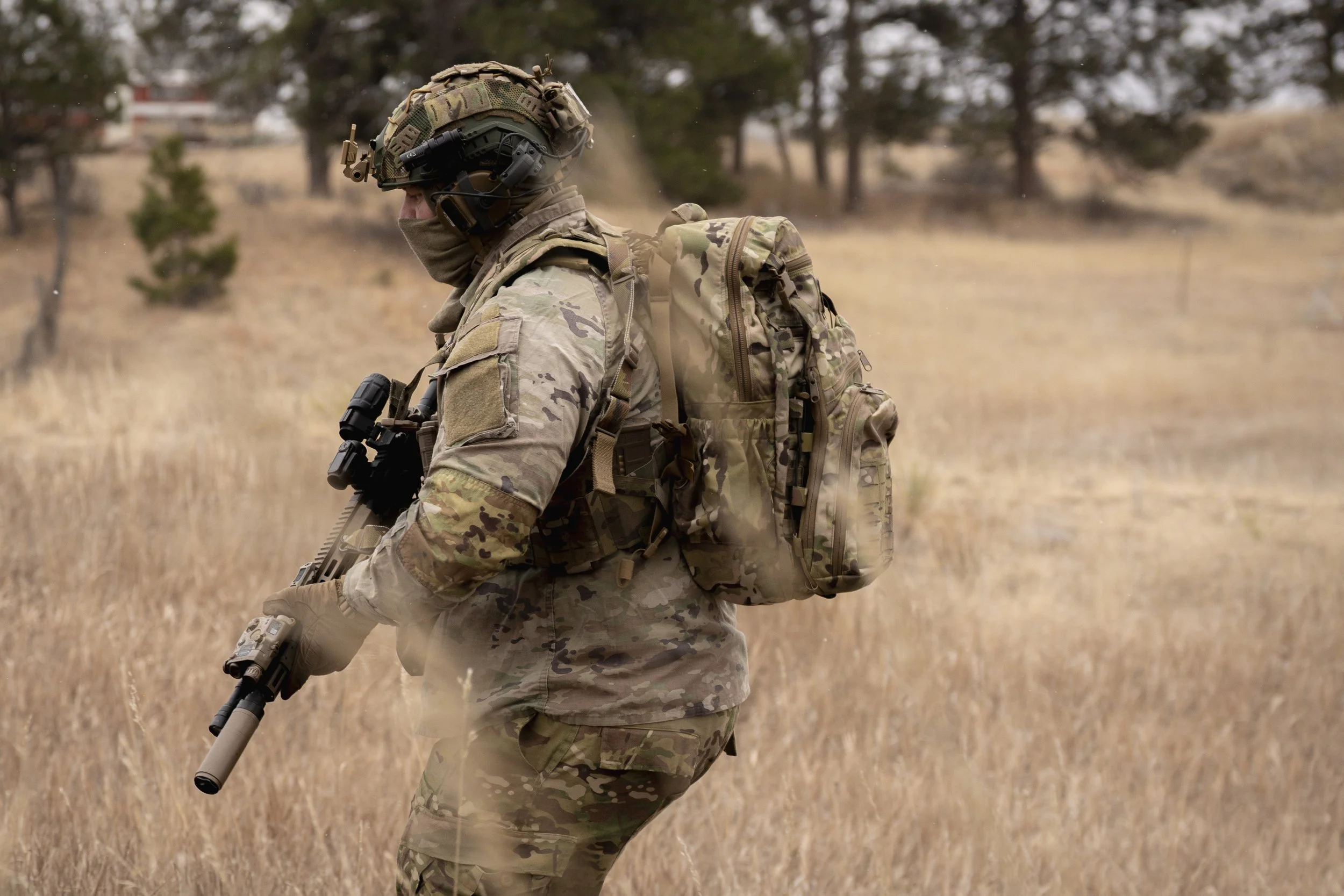 A soldier in camouflage uniform walking through a grassy field while holding a rifle, with trees in the background.
