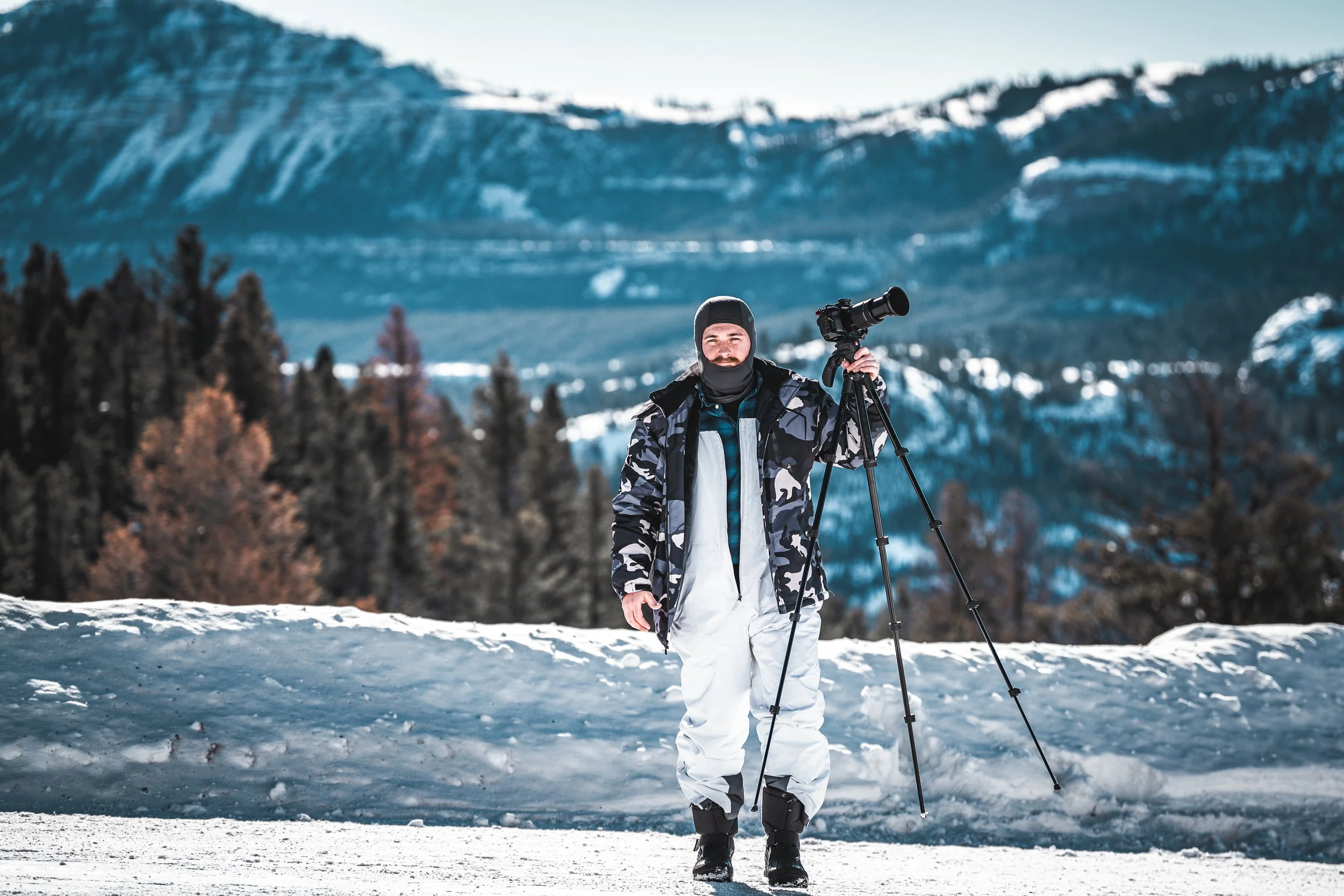 Man standing outdoors in snowy landscape with mountains and forest in background, holding a camera on a tripod.
