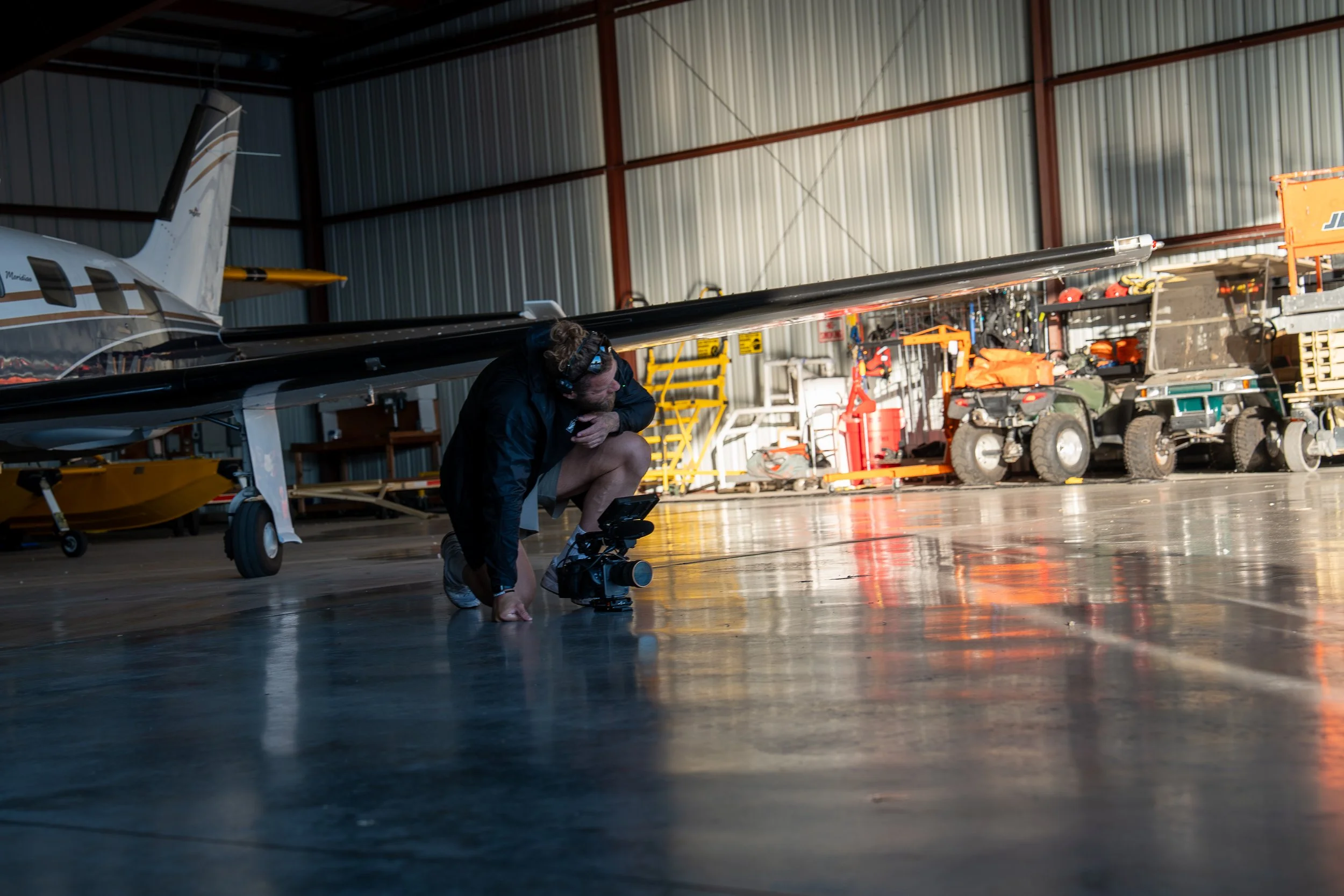A man crouches on the floor of an airplane hangar, adjusting equipment, with an airplane partially visible on the left and maintenance tools and vehicles in the background.