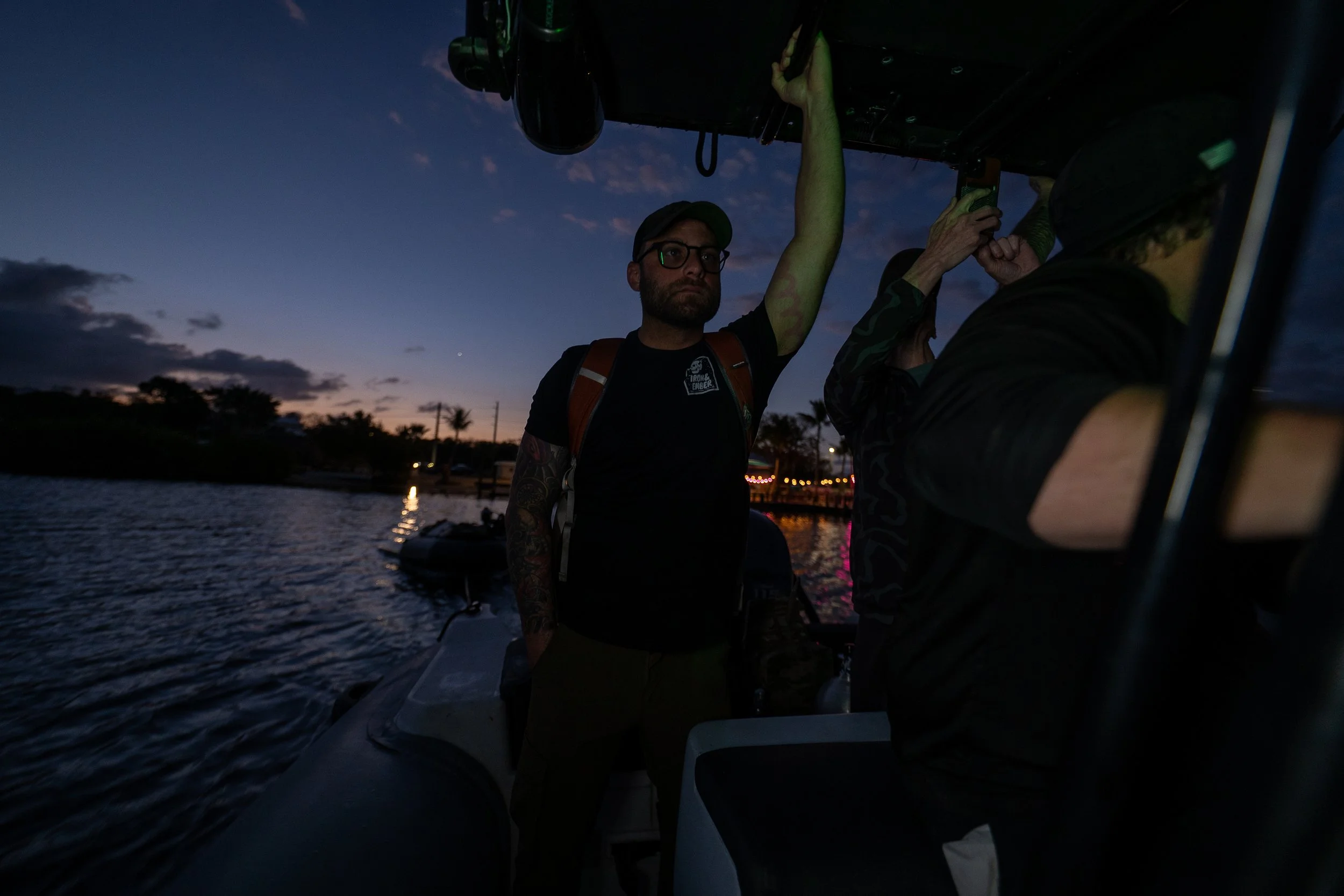 Two men on a boat at dusk, one with glasses and tattoos, holding onto the boat's structure, with a waterfront and palm trees in the background.