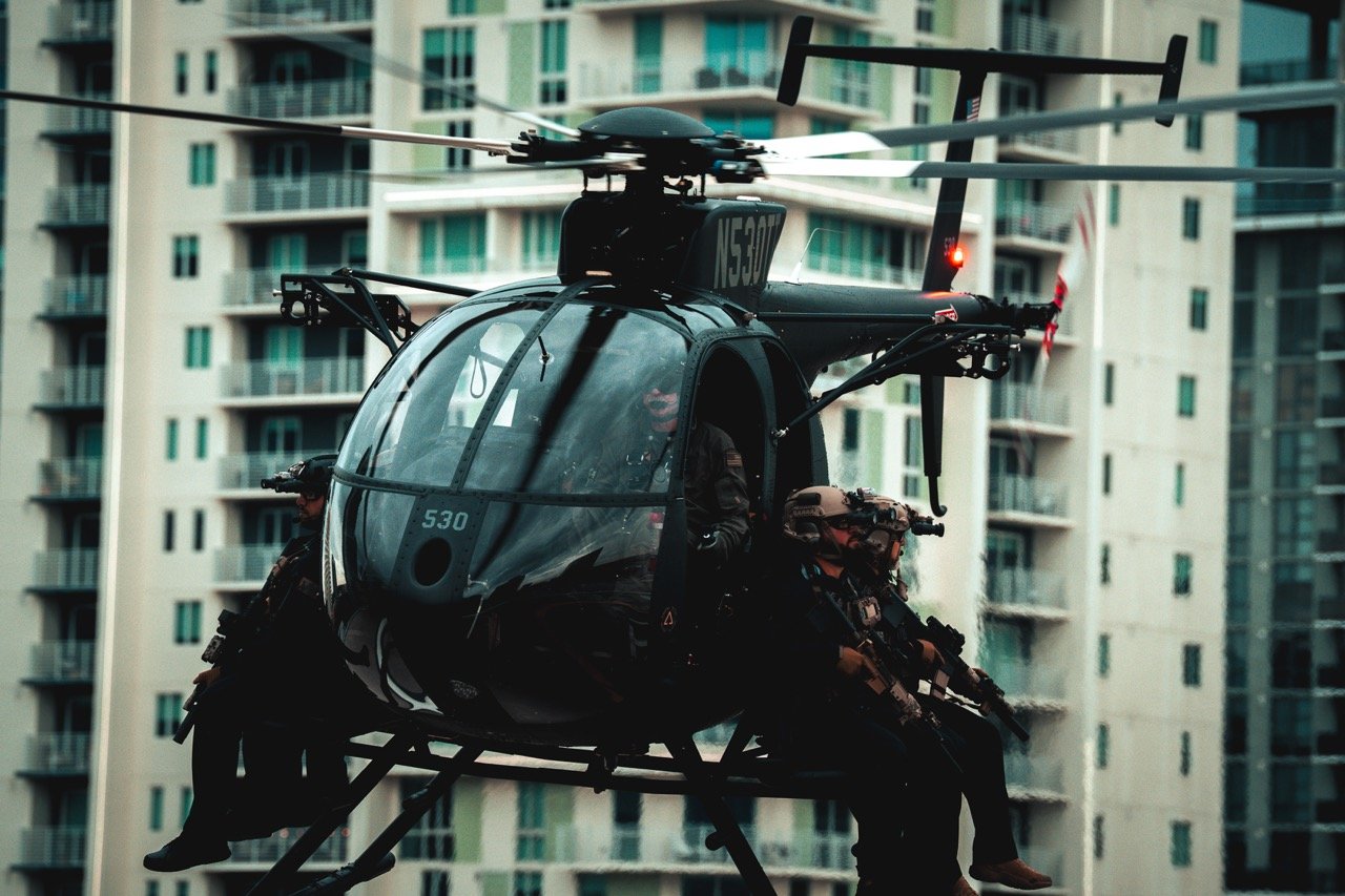 A black helicopter flying in front of tall residential buildings, with two soldiers onboard, armed and wearing tactical gear.