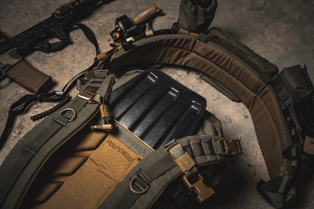 Tactical gear laid out on the ground, including a rifle, a magazine, a chest rig with a black plastic container, and a brown backpack.