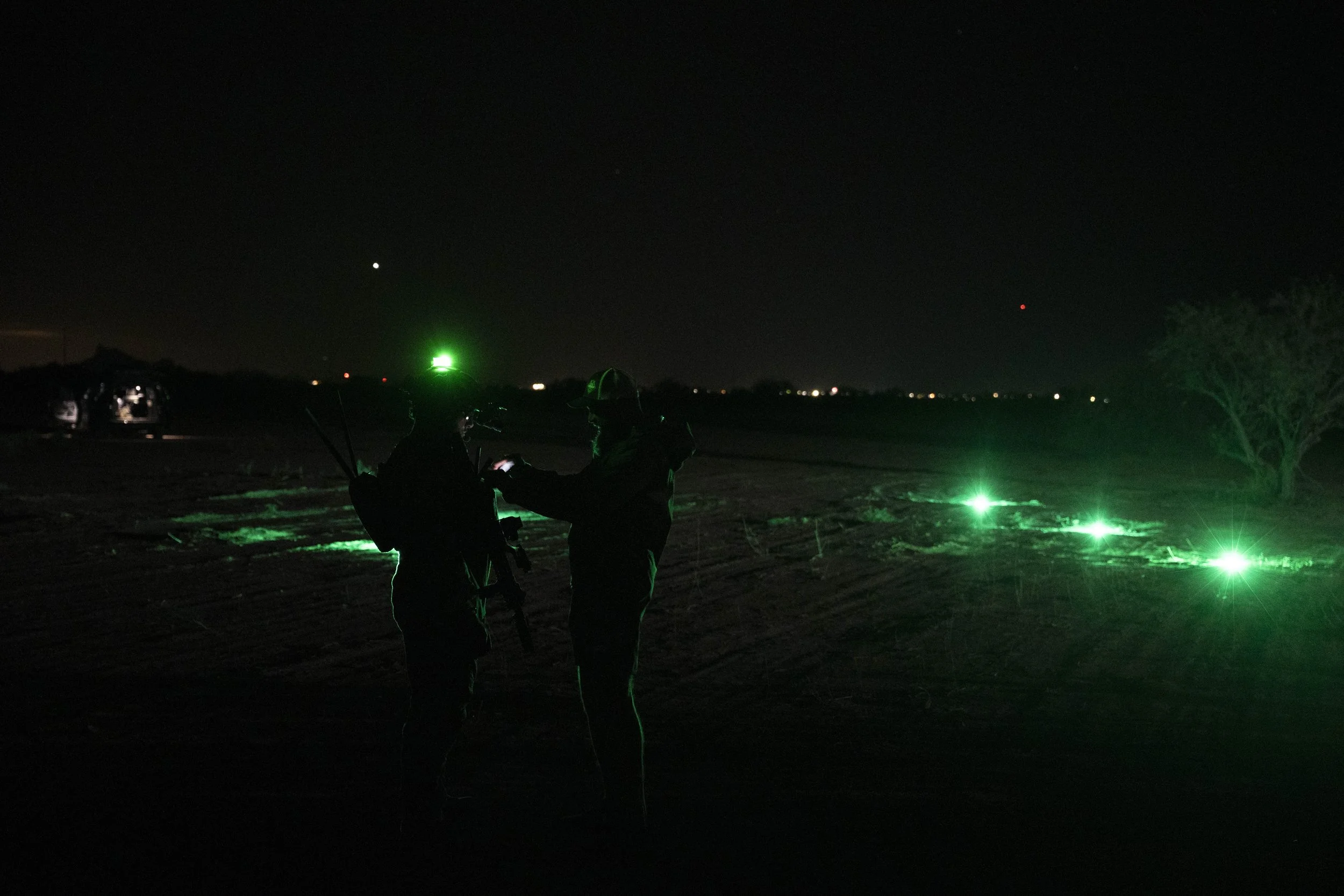Two silhouetted people in safety gear standing in a dark outdoor area, illuminated by green lights, with a tree on the right and a distant horizon with faint city lights.