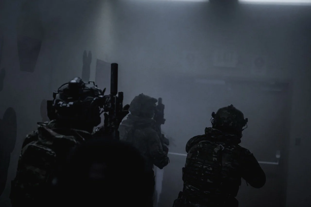 Three soldiers in combat gear moving through a dark, smoky environment, potentially during a military operation.