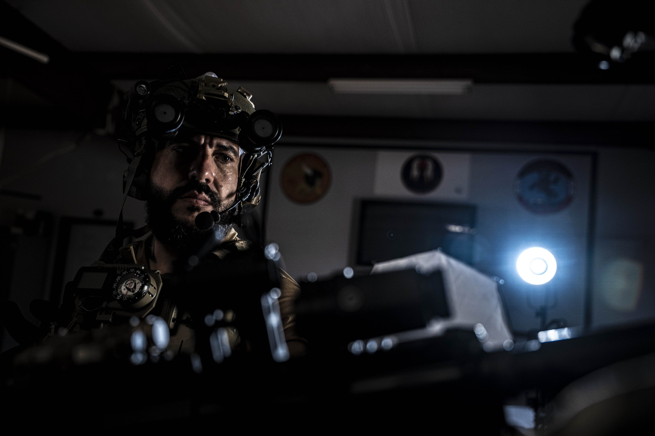 A man wearing military gear and night vision equipment, sitting in a dimly lit room.