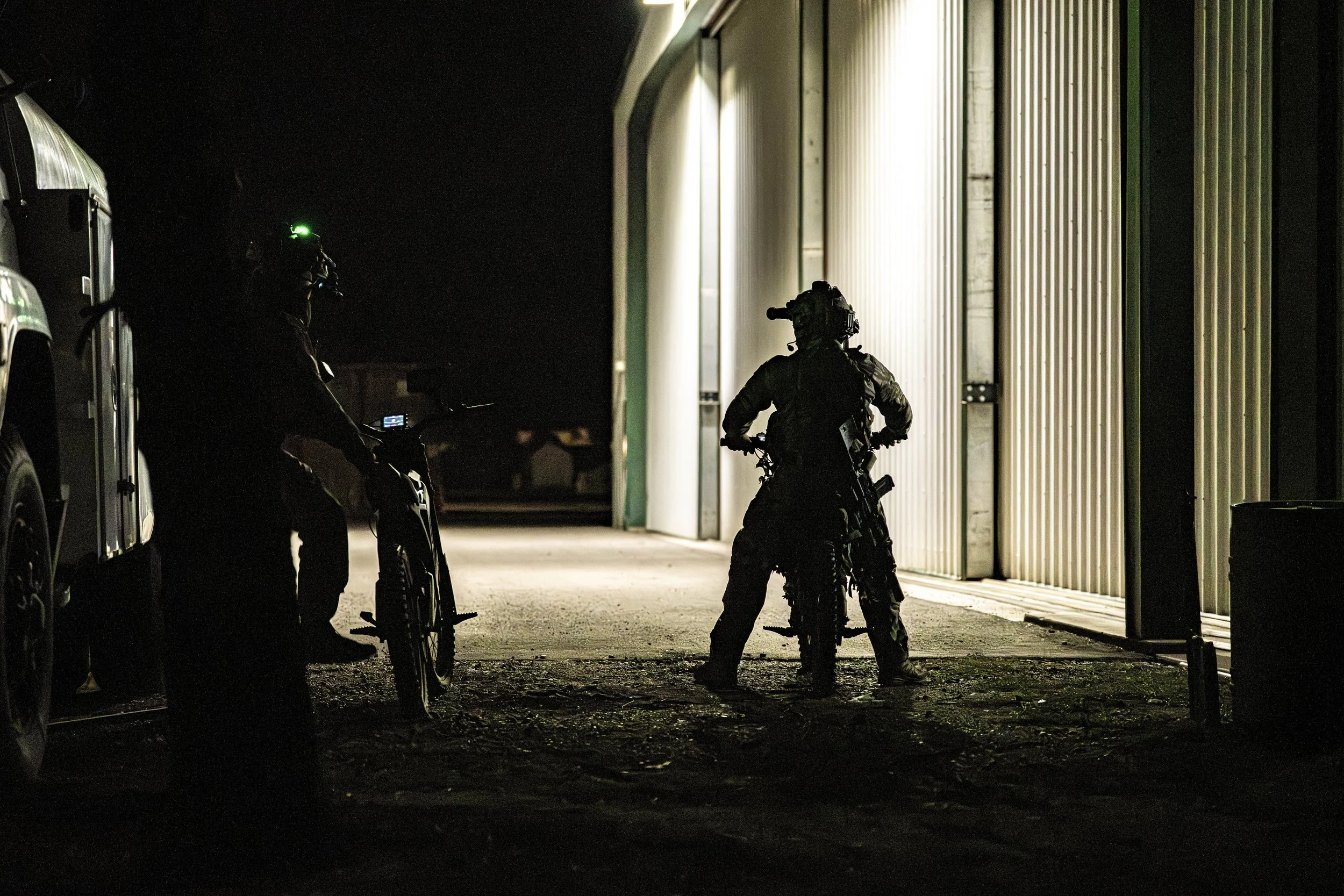 Silhouettes of police officers wearing tactical gear at night near a warehouse, one on a bike and another standing in front of a large open door, illuminated by external light.