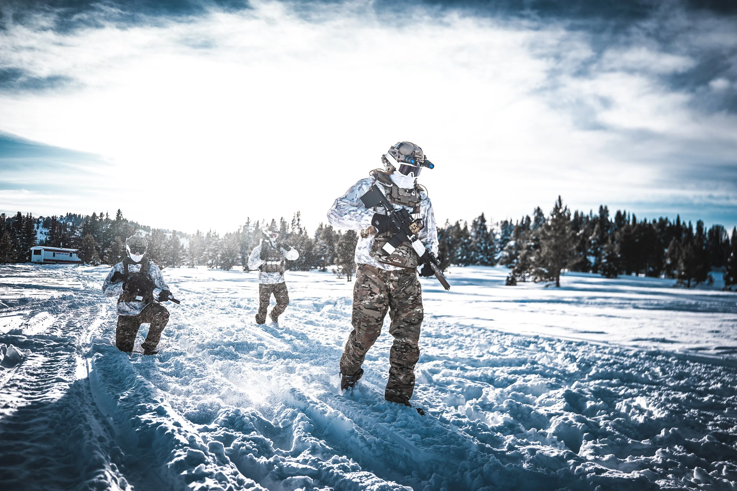 Three soldiers in camouflage winter gear and tactical equipment walking through snowy terrain with a forest and cloudy sky in the background.