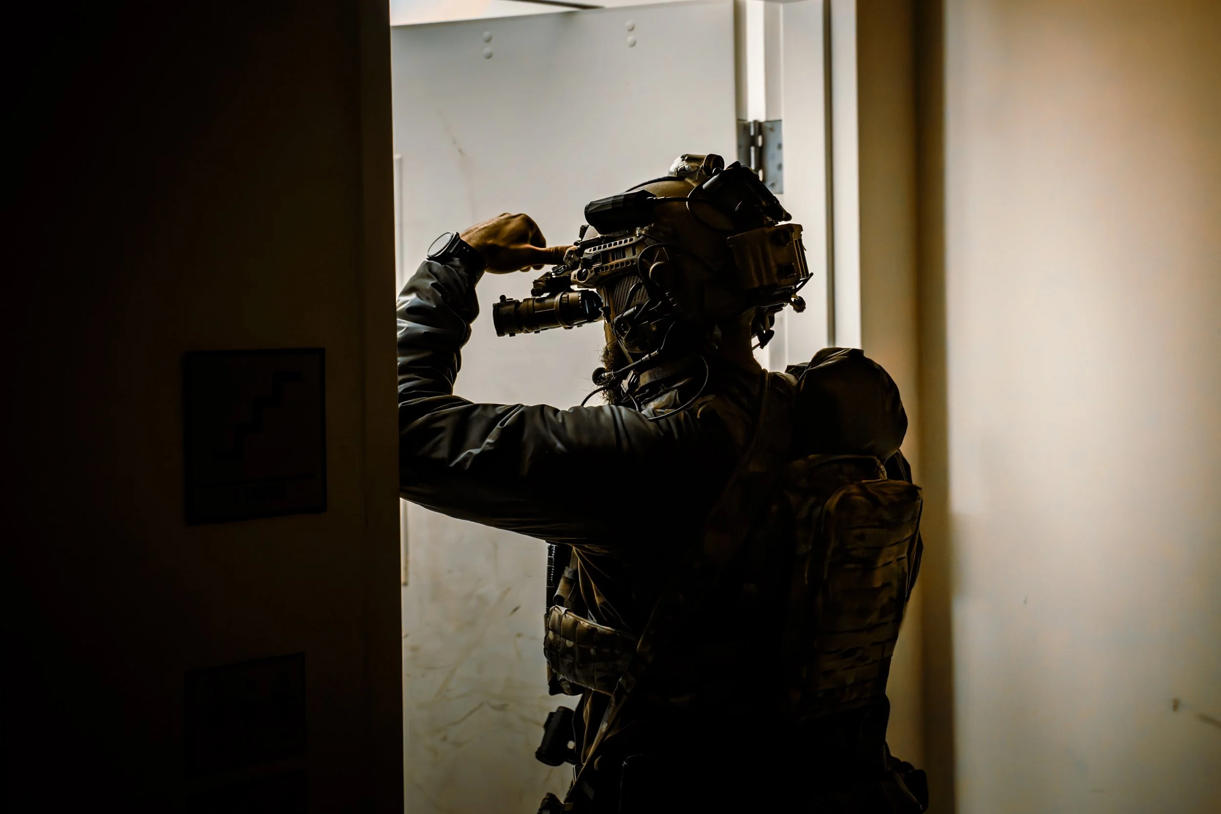 Military personnel in tactical gear, silhouette, aiming a firearm while looking through the sights, partially hidden behind a door.