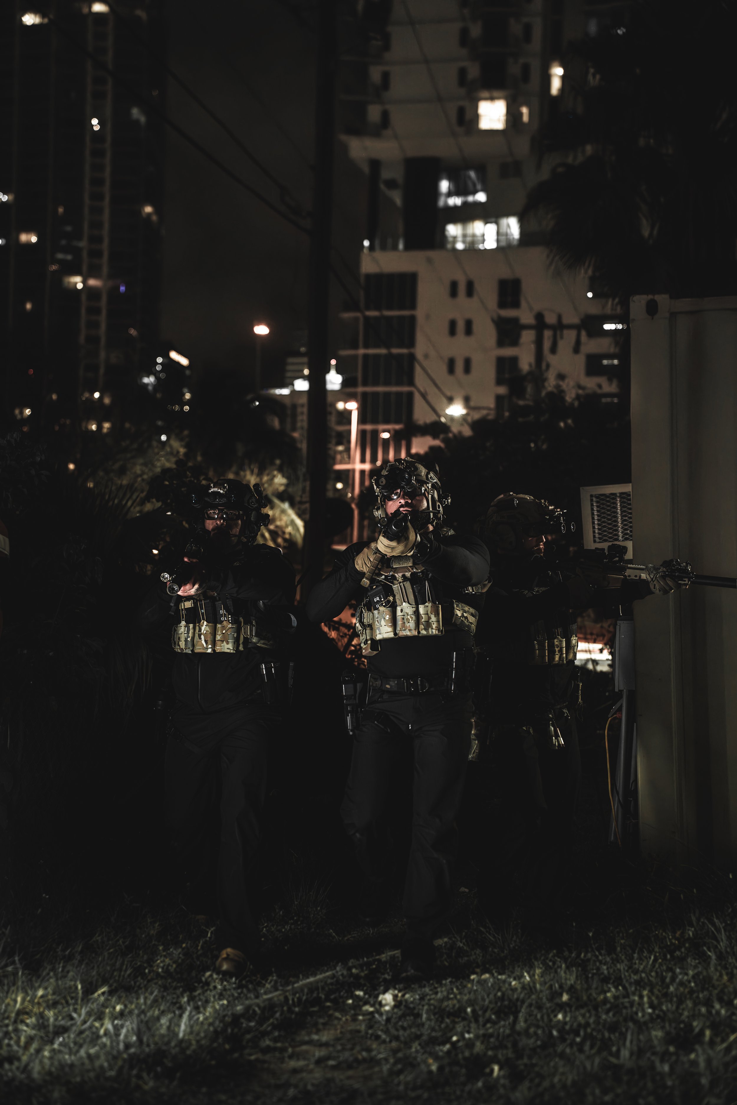 Three police officers in tactical gear at night, holding weapons, with city buildings in the background.