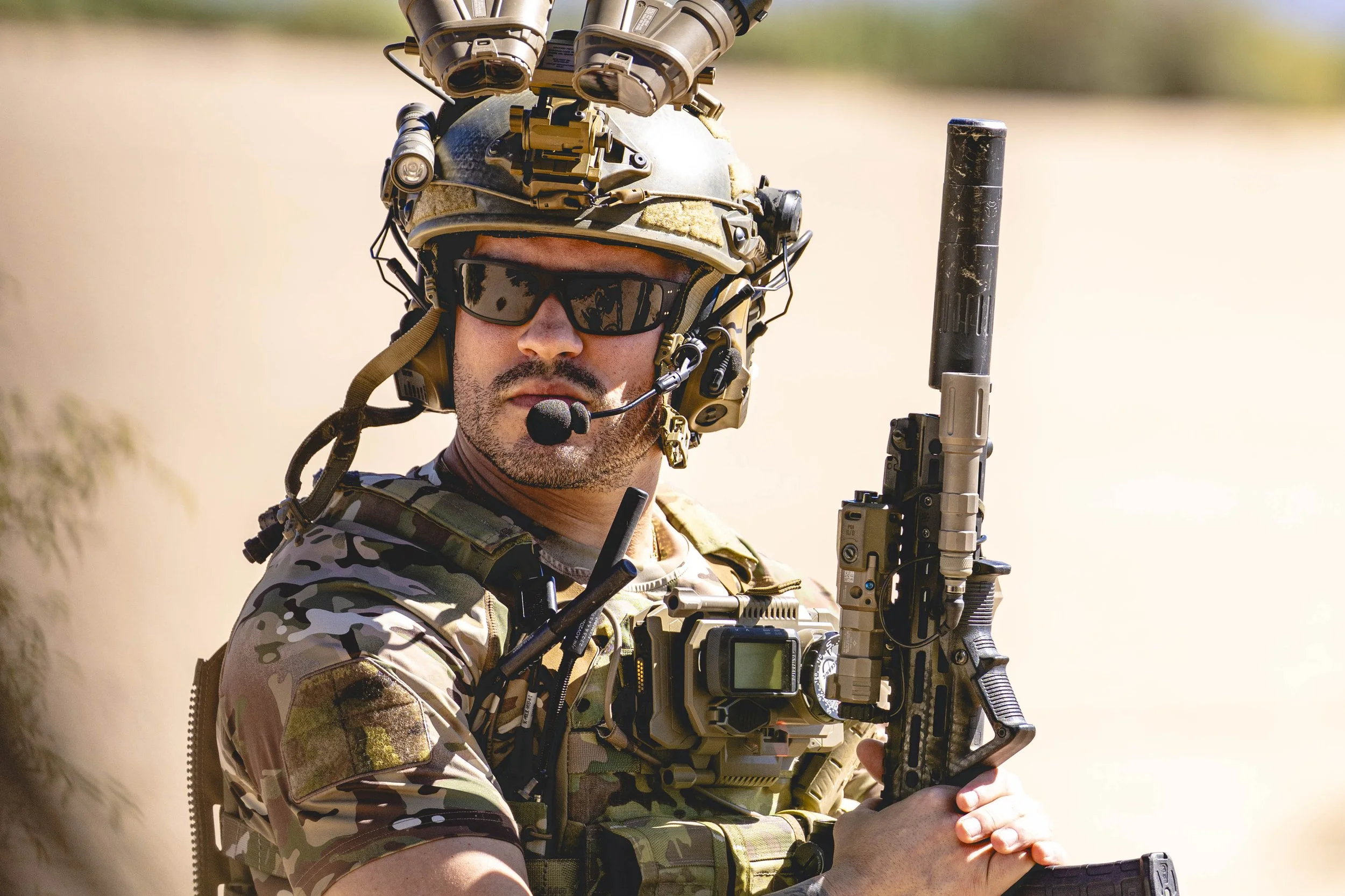 A soldier in camouflage uniform and tactical gear, wearing sunglasses, a helmet with mounted equipment, and holding a rifle with a silencer.
