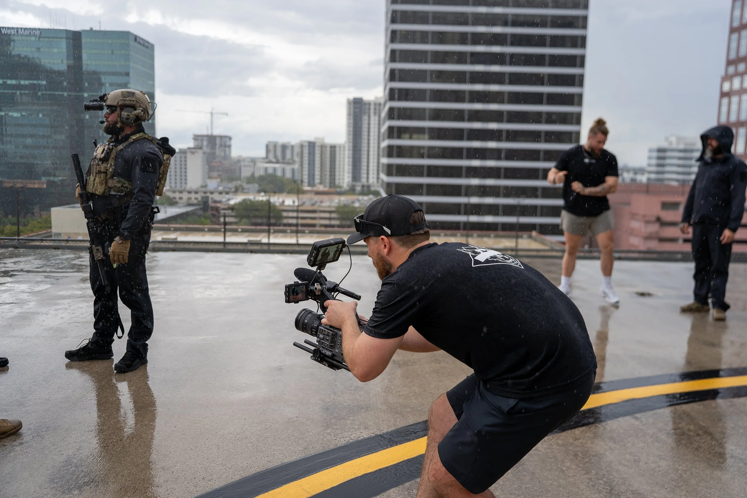 A group of people, including a cameraman filming, on a rainy rooftop in an urban area with high-rise buildings in the background.