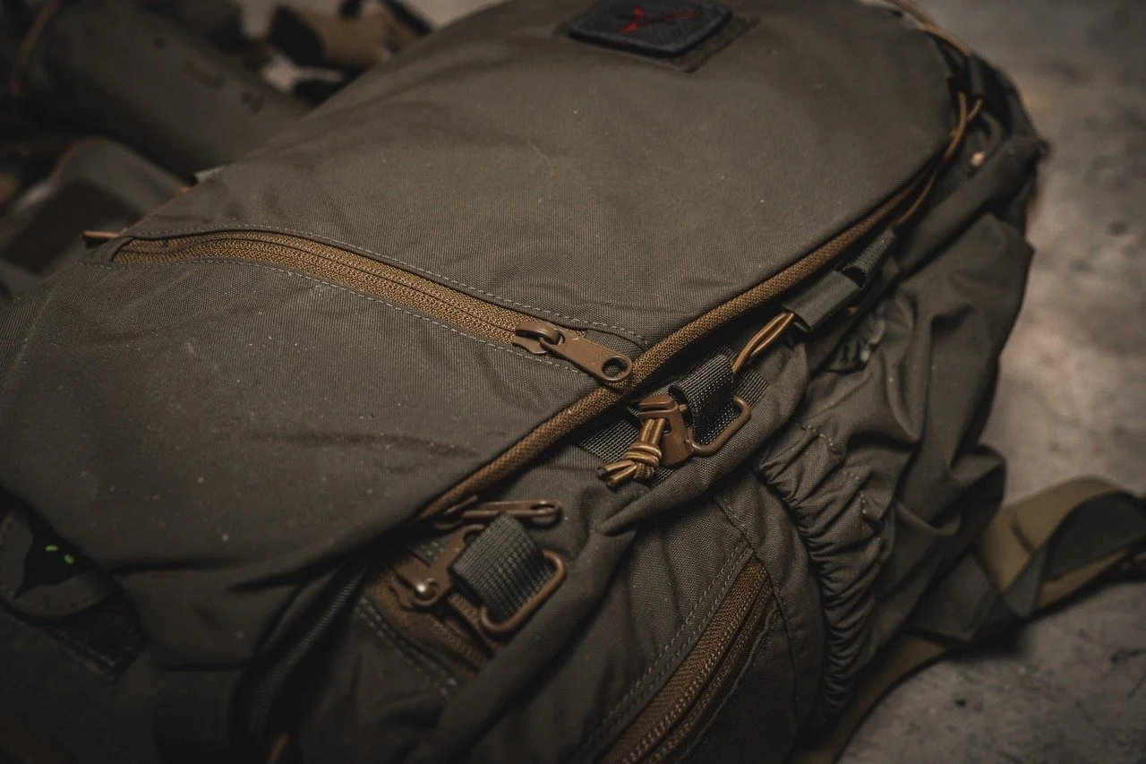 A dark brown or black tactical or travel backpack with multiple zippered compartments and metal buckles.