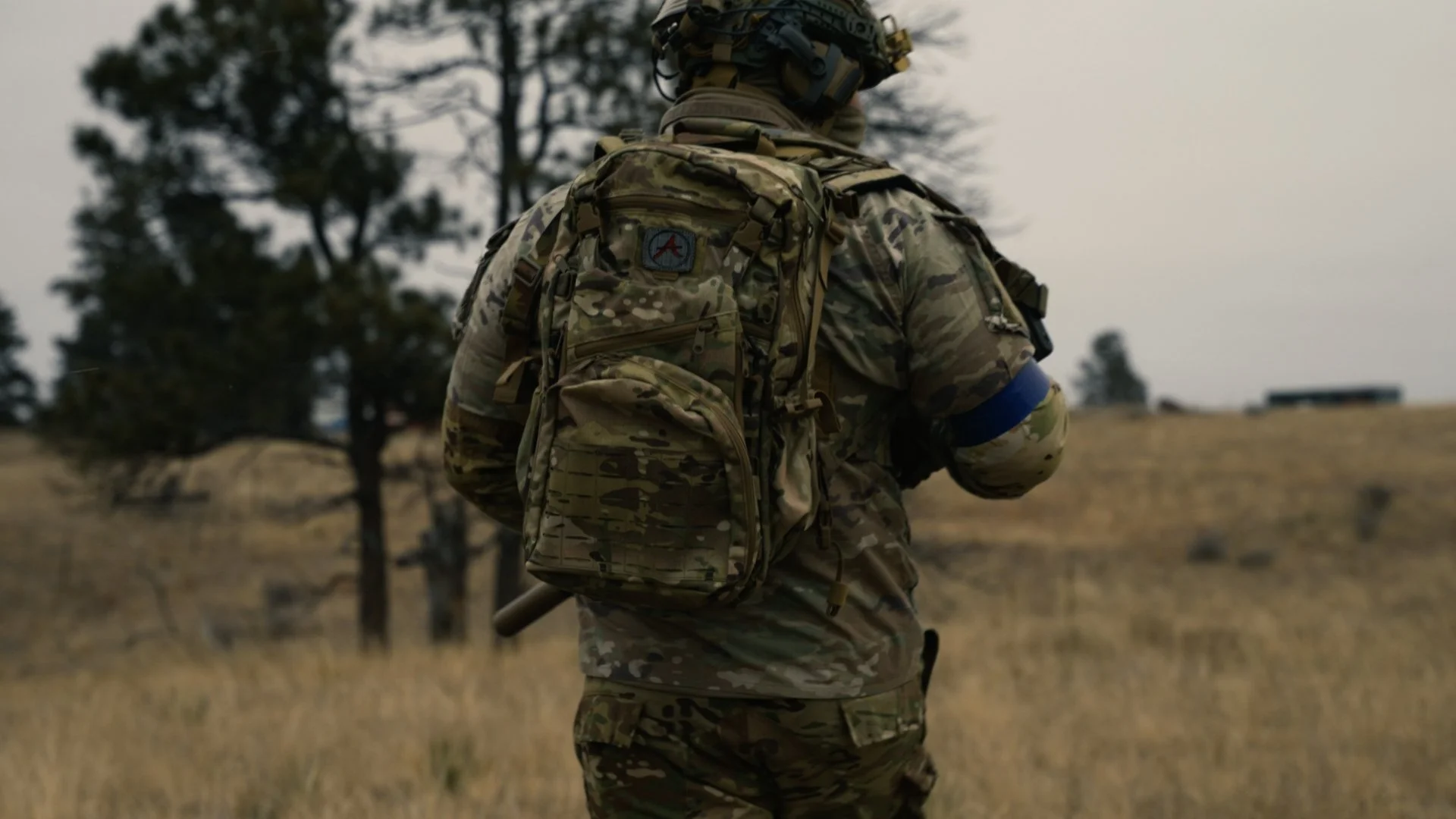 A soldier in camouflage uniform with a large tactical backpack walking through a field with trees in the background.