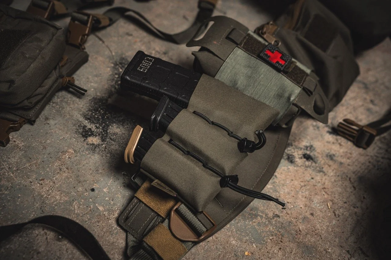 Military or tactical gear on a concrete floor, including a green pouch, a black magazine pouch, and a first aid kit with a red cross.