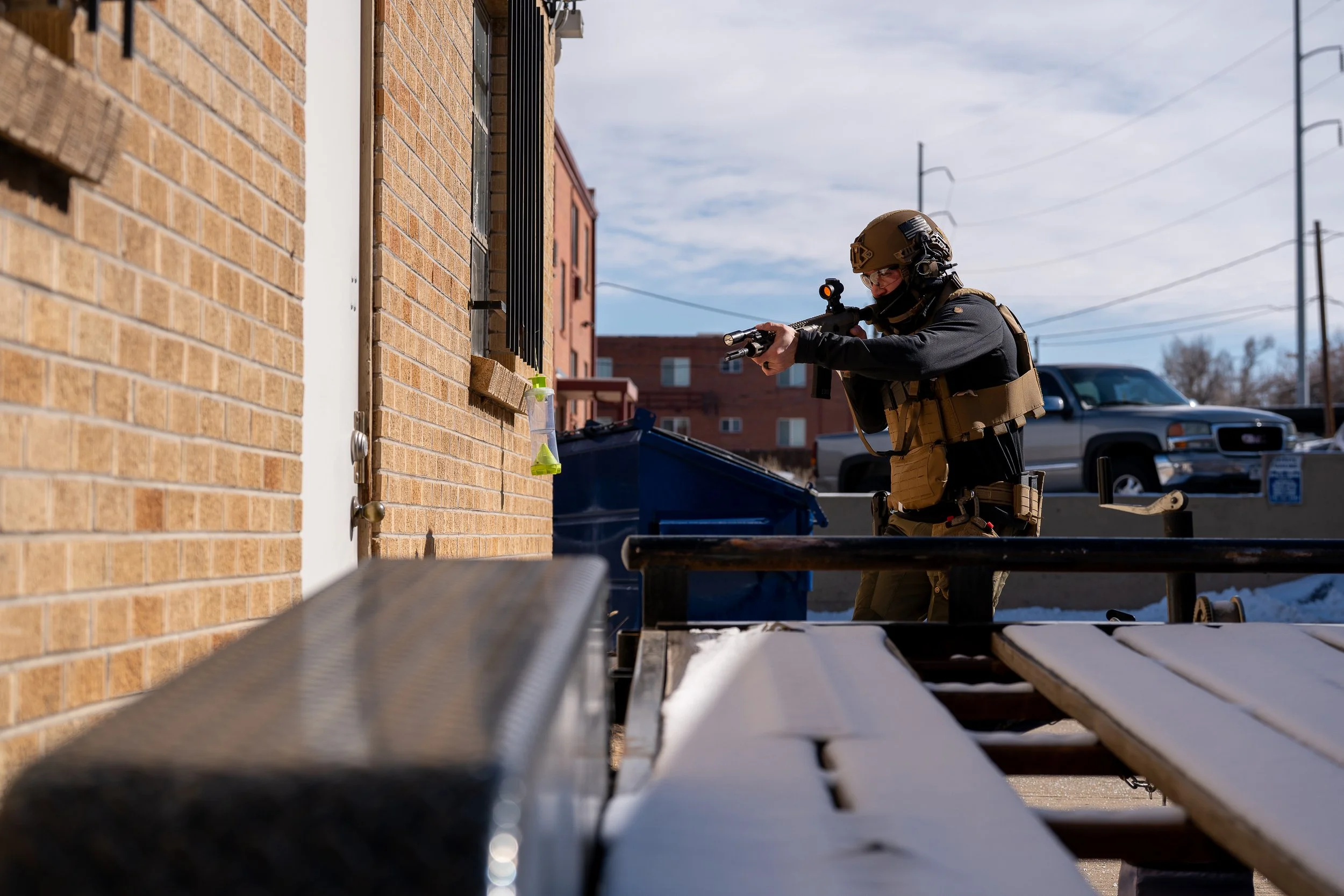 A law enforcement officer aiming a rifle outside a brick building on a city street.