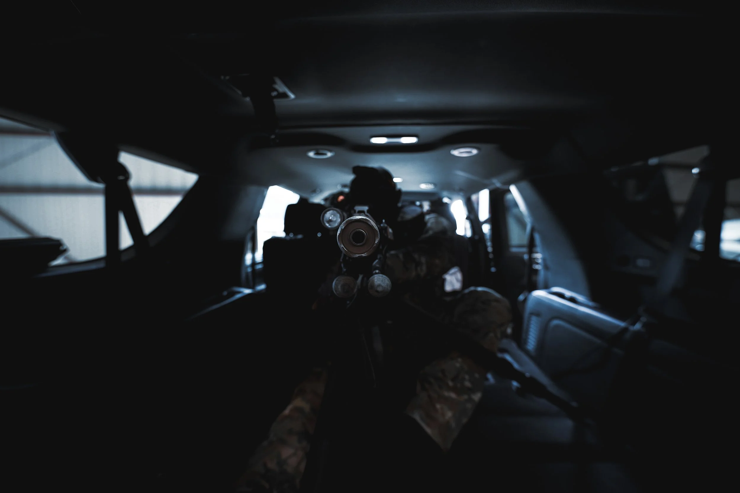 Soldier aiming weapon from the back of military vehicle inside a dimly lit interior.