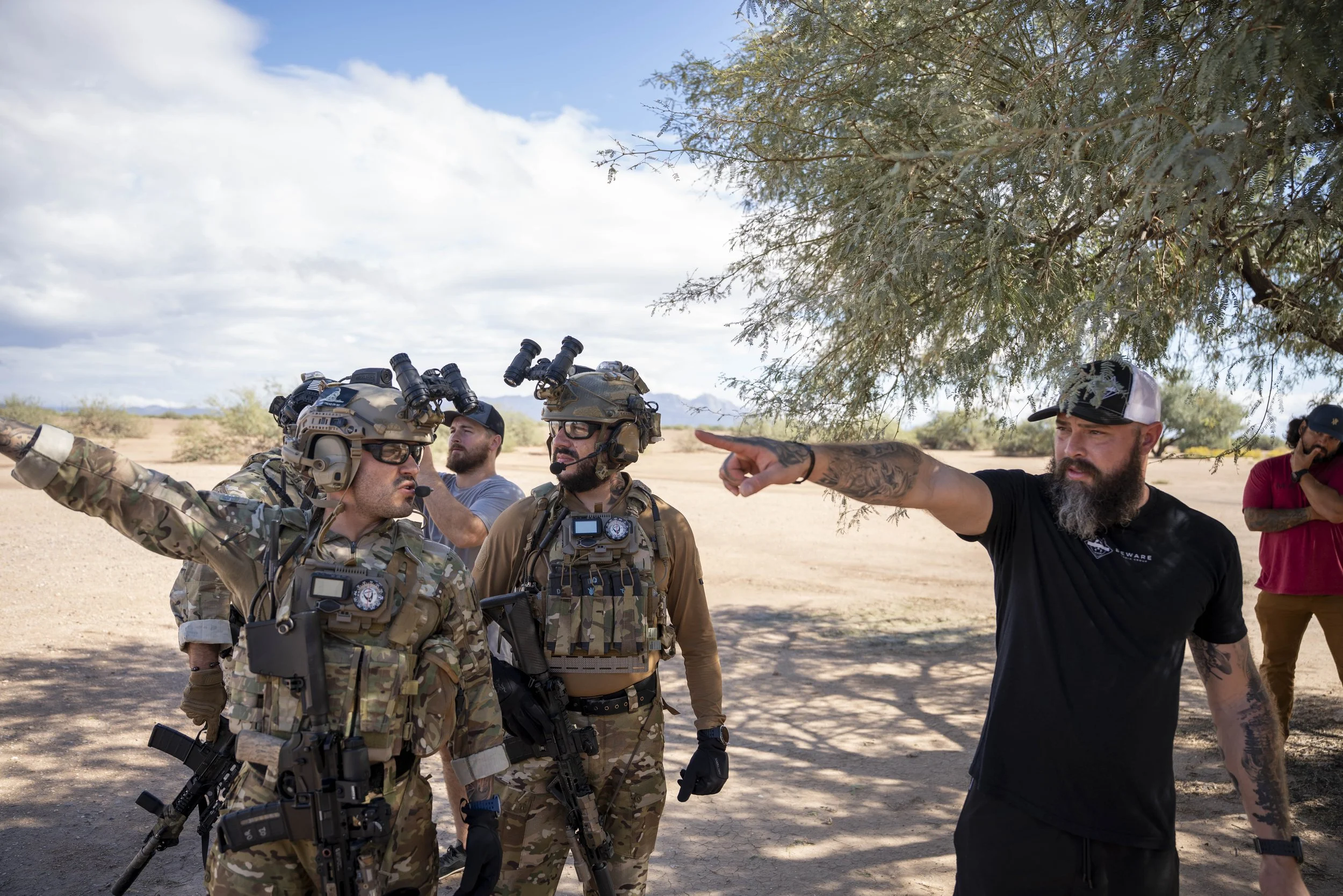 Military personnel in camouflage uniforms and tactical gear standing under a tree, receiving instructions from a man in a black T-shirt with a beard, while others stand in the background in a desert landscape under a partly cloudy sky.