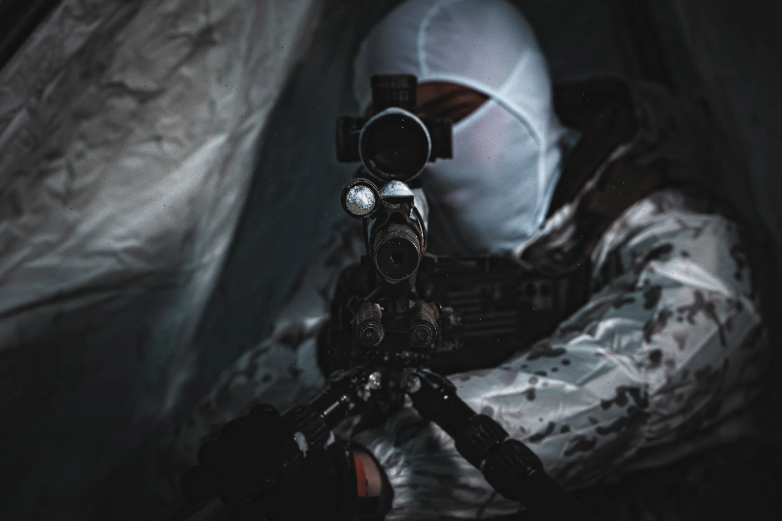 Person in a white camouflage suit aiming a sniper rifle inside a tent or small space.
