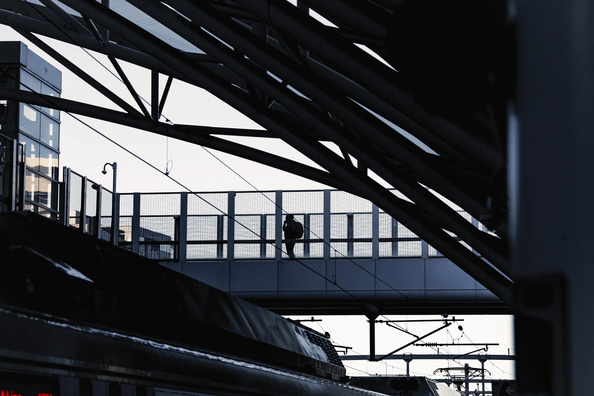 Silhouette of a person with a backpack walking on an elevated pedestrian walkway in a train station with overhead electrical lines and modern building in the background.