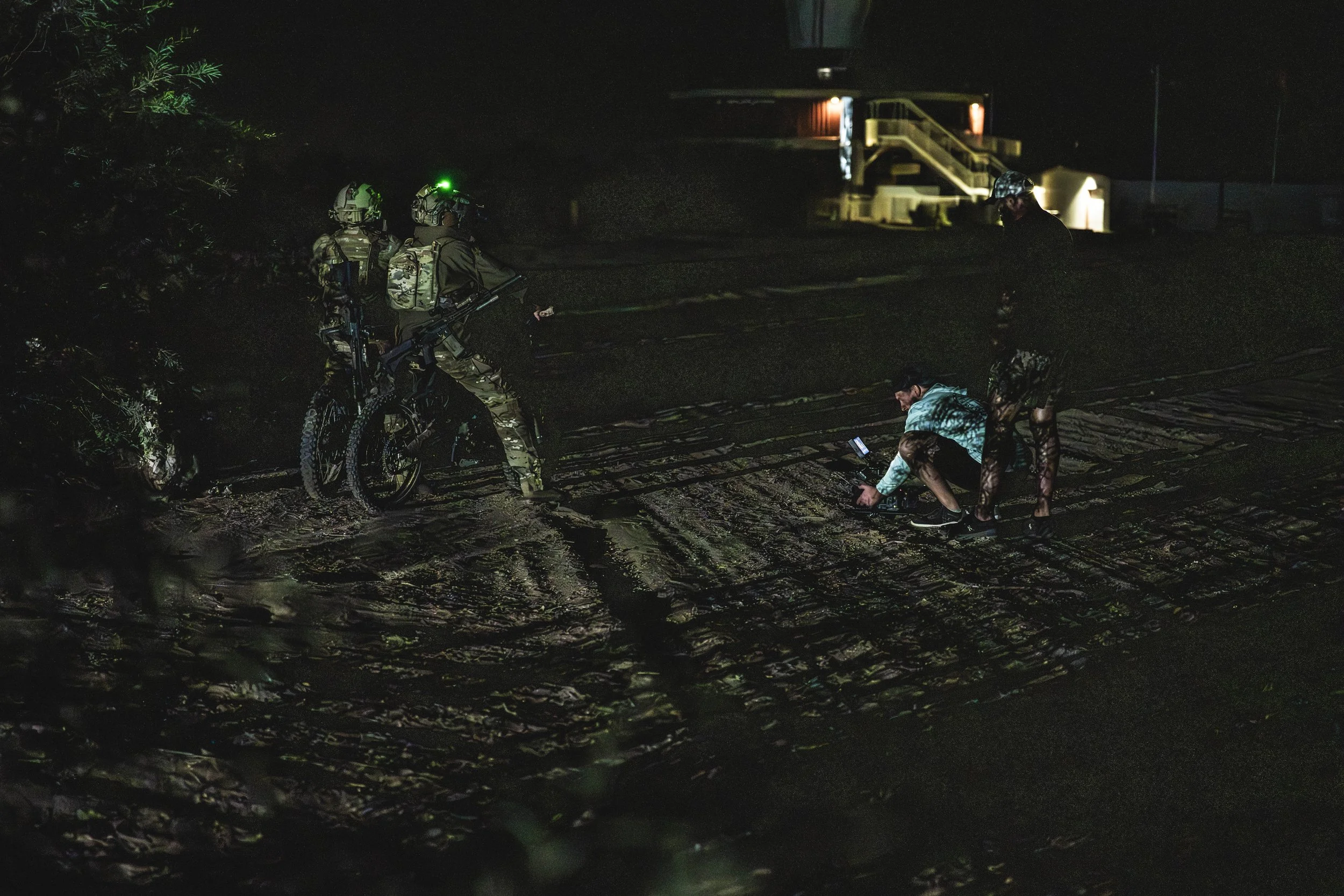 Nighttime scene with two soldiers in camouflage uniforms and helmets, one on a bicycle with a green light on the helmet, and one person crouching on the ground with a phone, all on a muddy area near a house or building with stairs and lights in the b