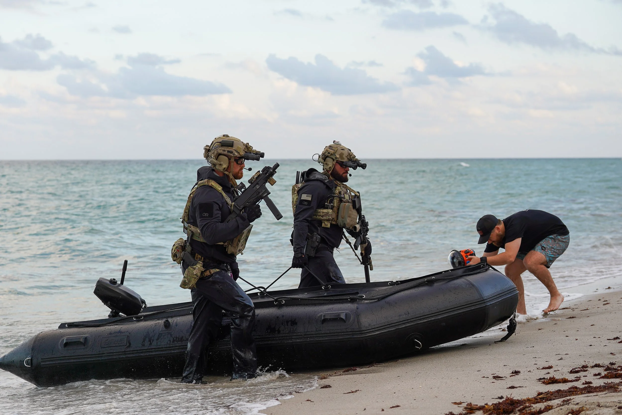 Two soldiers in tactical gear and helmets with night vision goggles board a black inflatable boat on a beach, while a man in casual clothes adjusts a device on the boat