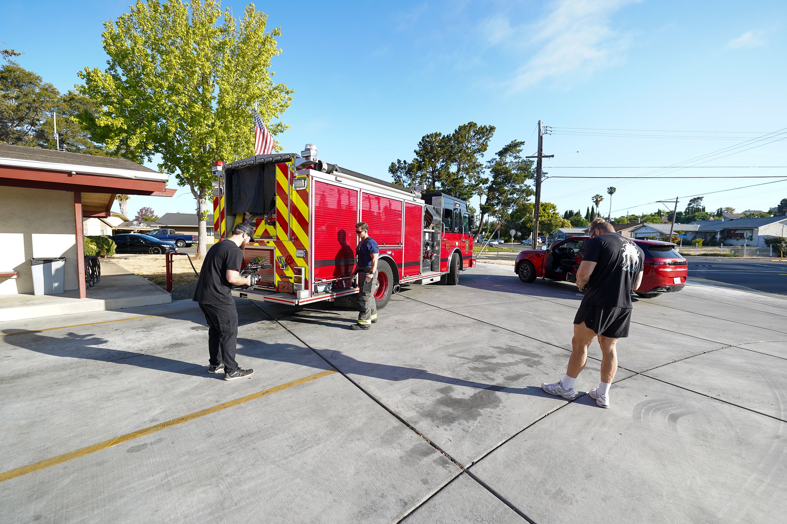 Firefighters preparing equipment next to a red fire truck on a driveway, with a person standing nearby and a red car with open doors in the background on a sunny day.