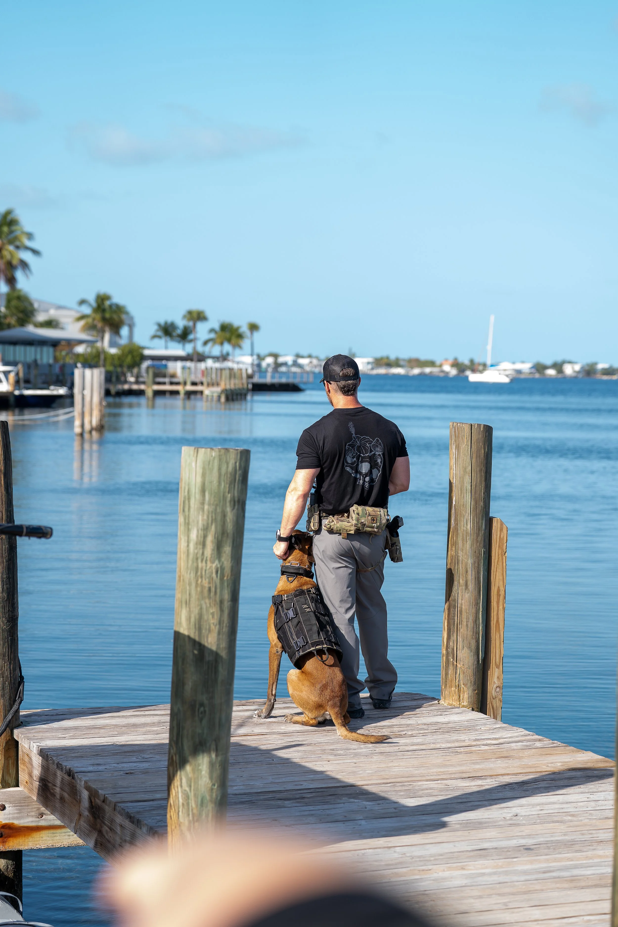 A man with a police dog on a wooden dock overlooking a waterway with houses and palms in the background.