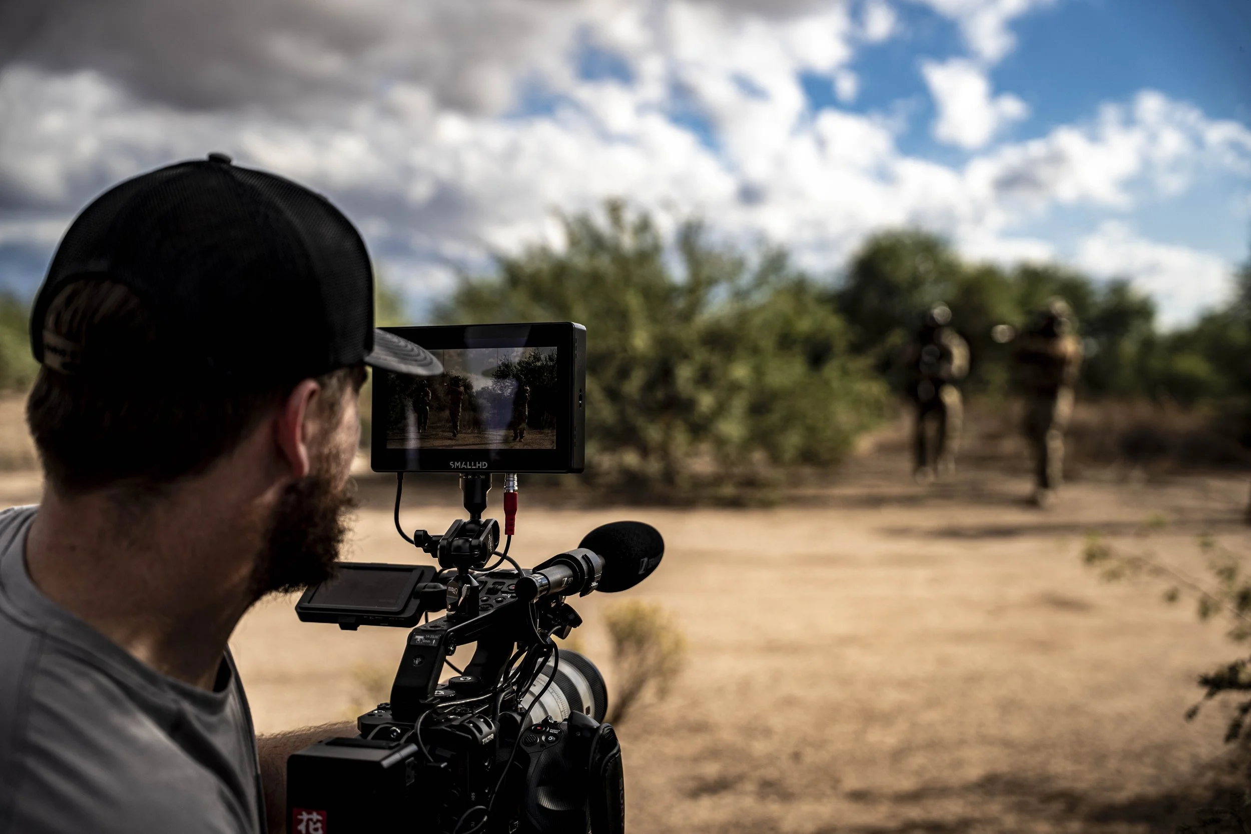 A man wearing a black cap operates a professional video camera, filming two people walking in a desert landscape with bushes and a partly cloudy sky.
