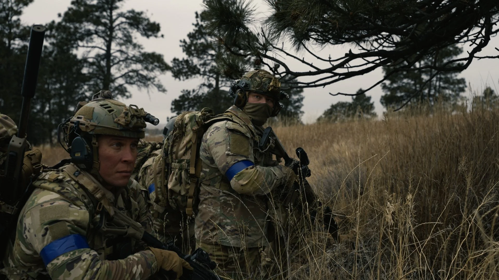 Two soldiers in camouflage uniforms and helmets, with blue armbands, crouching in a grassy field with trees in the background.