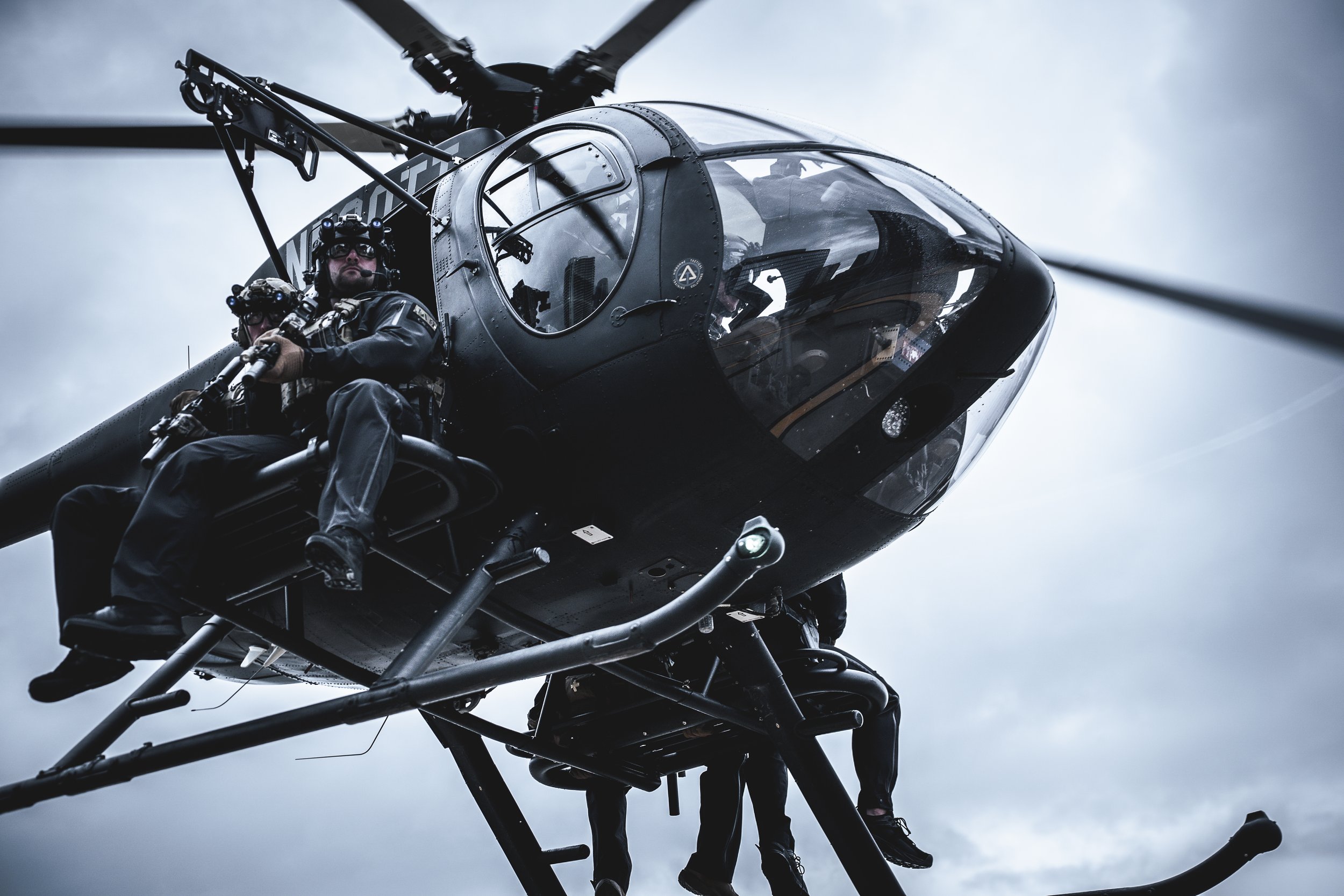 A black helicopter with two crew members sitting outside, wearing helmets and gear, flying against a cloudy sky.