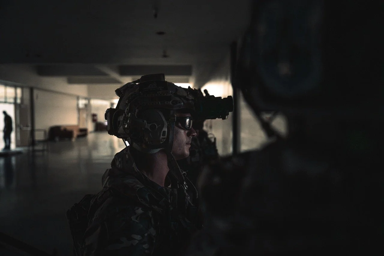 A soldier wearing night vision gear and camouflage uniform in a dark indoor setting.