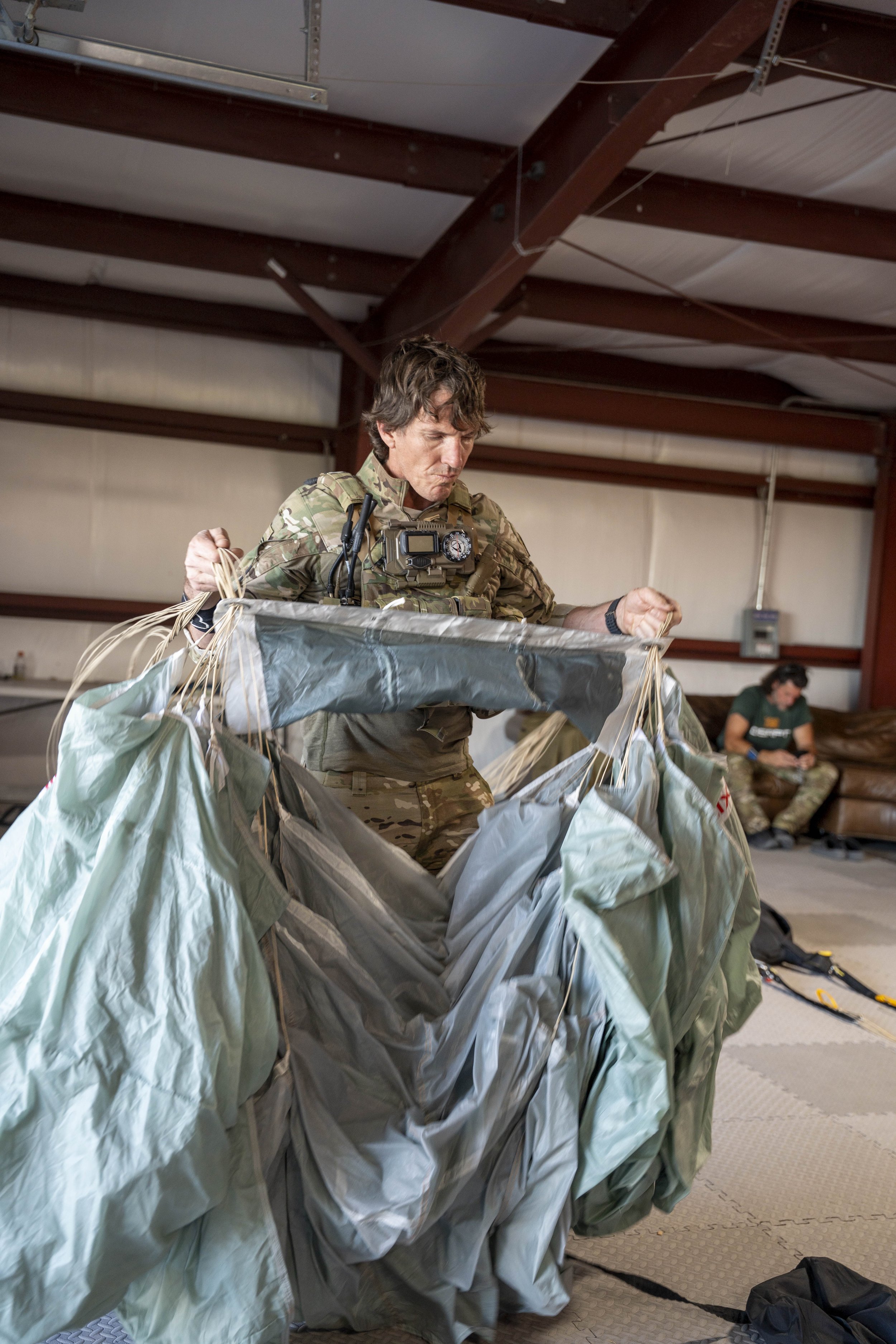 A soldier wearing camouflage uniform is preparing to skydive, holding onto the edges of an open parachute inside a hangar or training facility. In the background, a woman in military attire is sitting on a couch, looking at her phone.