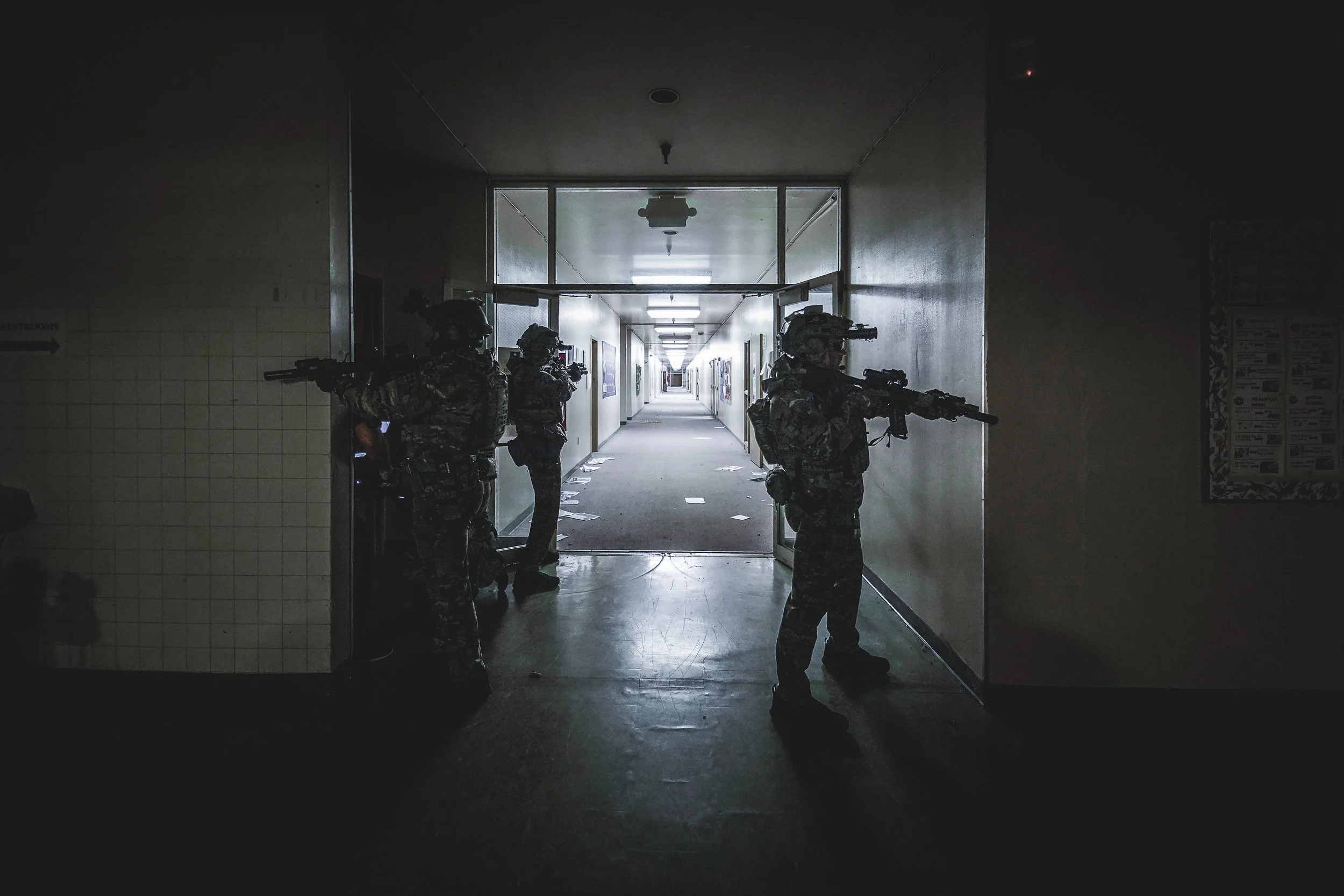 Four soldiers in camouflage gear and helmets armed with rifles taking position in a dimly lit hallway.