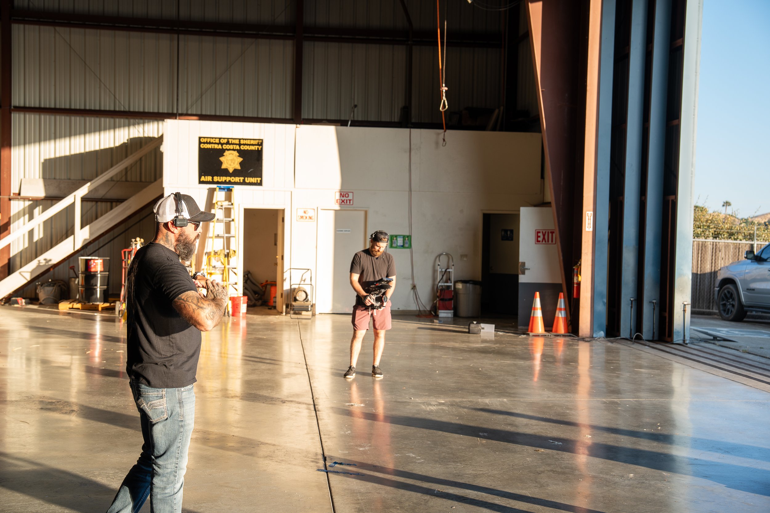 Two men inside a warehouse, one with a headset and sunglasses looking to the left, and another holding a camera in the background. The warehouse has signs, safety cones, and an open garage door revealing an outdoor area.