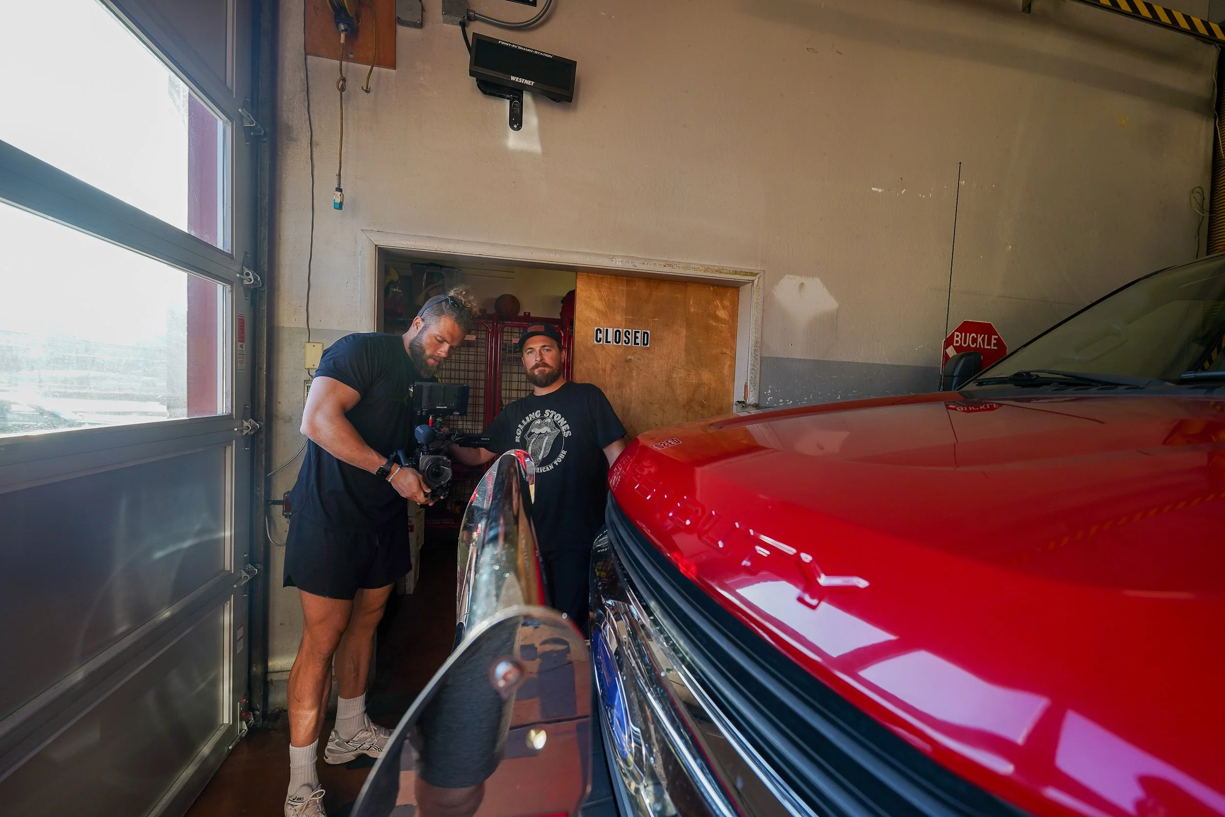 Two men standing inside a garage next to a red vehicle, with one man holding a camera and the other leaning against the door with a "CLOSED" sign, sunlight coming through a window on the left.