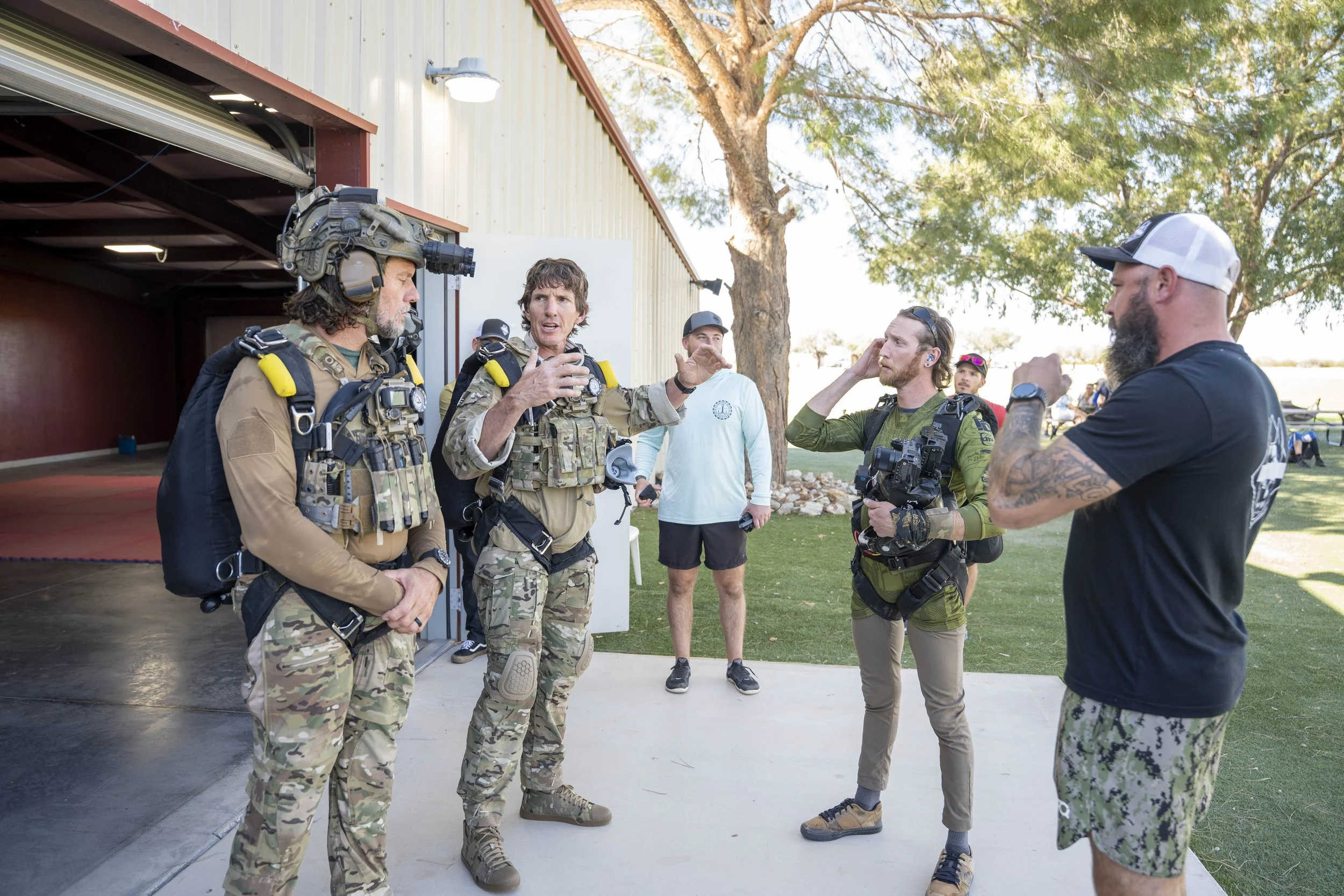Group of soldiers and camera crew having a discussion outdoors near an open garage, with a tree and grass in the background.