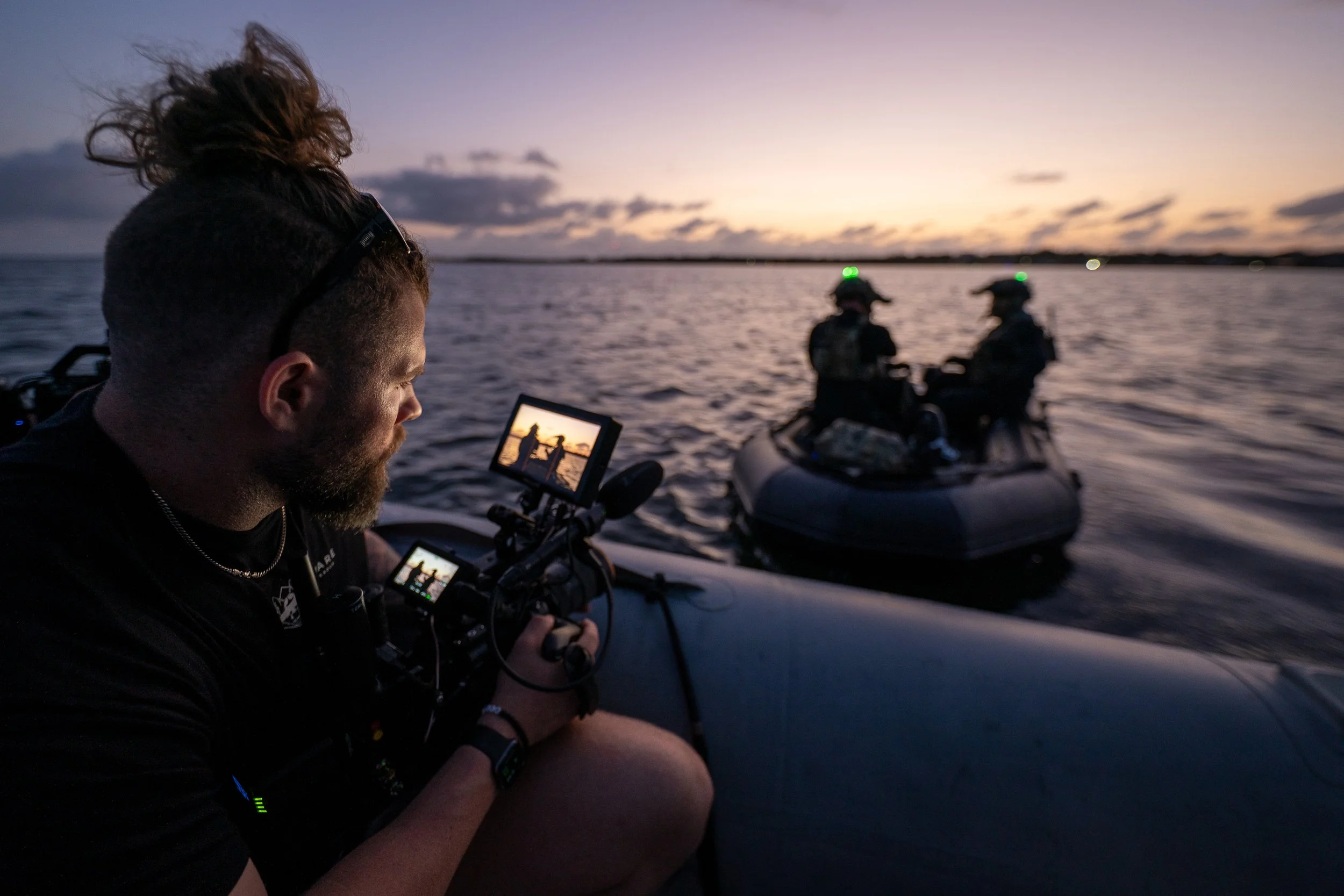 A man operating a camera on a boat during sunset, filming two soldiers in an inflatable boat on a body of water.