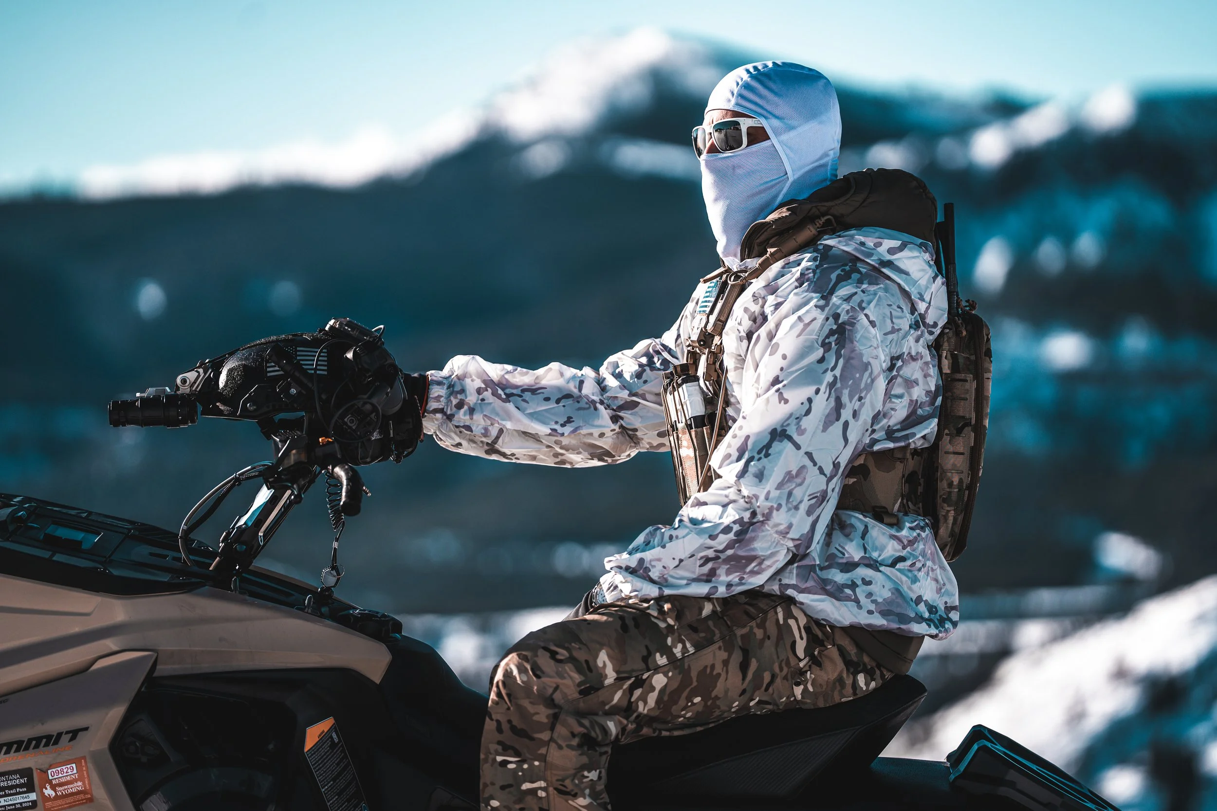 A person riding a snowmobile in a snowy landscape, dressed in camouflage and winter gear with a face covering and sunglasses.
