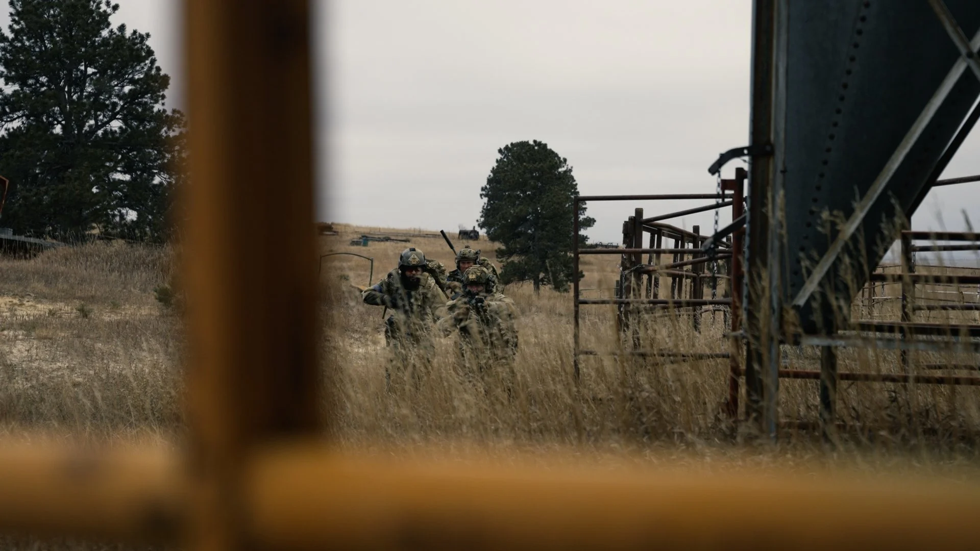 Three soldiers in camouflage gear walking through a grassy field, viewed through a yellow metal fence, with trees and overcast sky in the background.