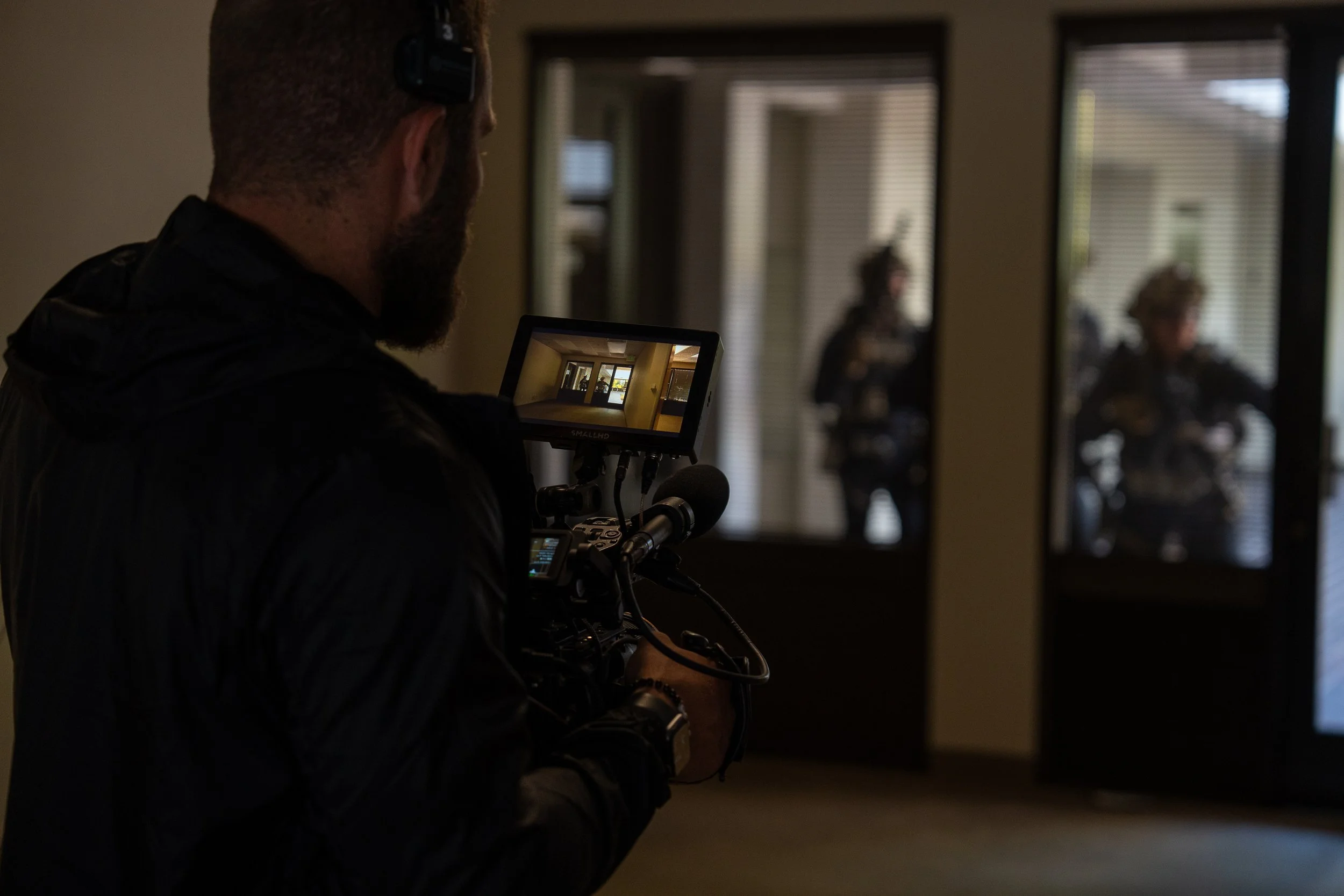 A cameraman filming a scene with two police officers standing outside through glass doors.