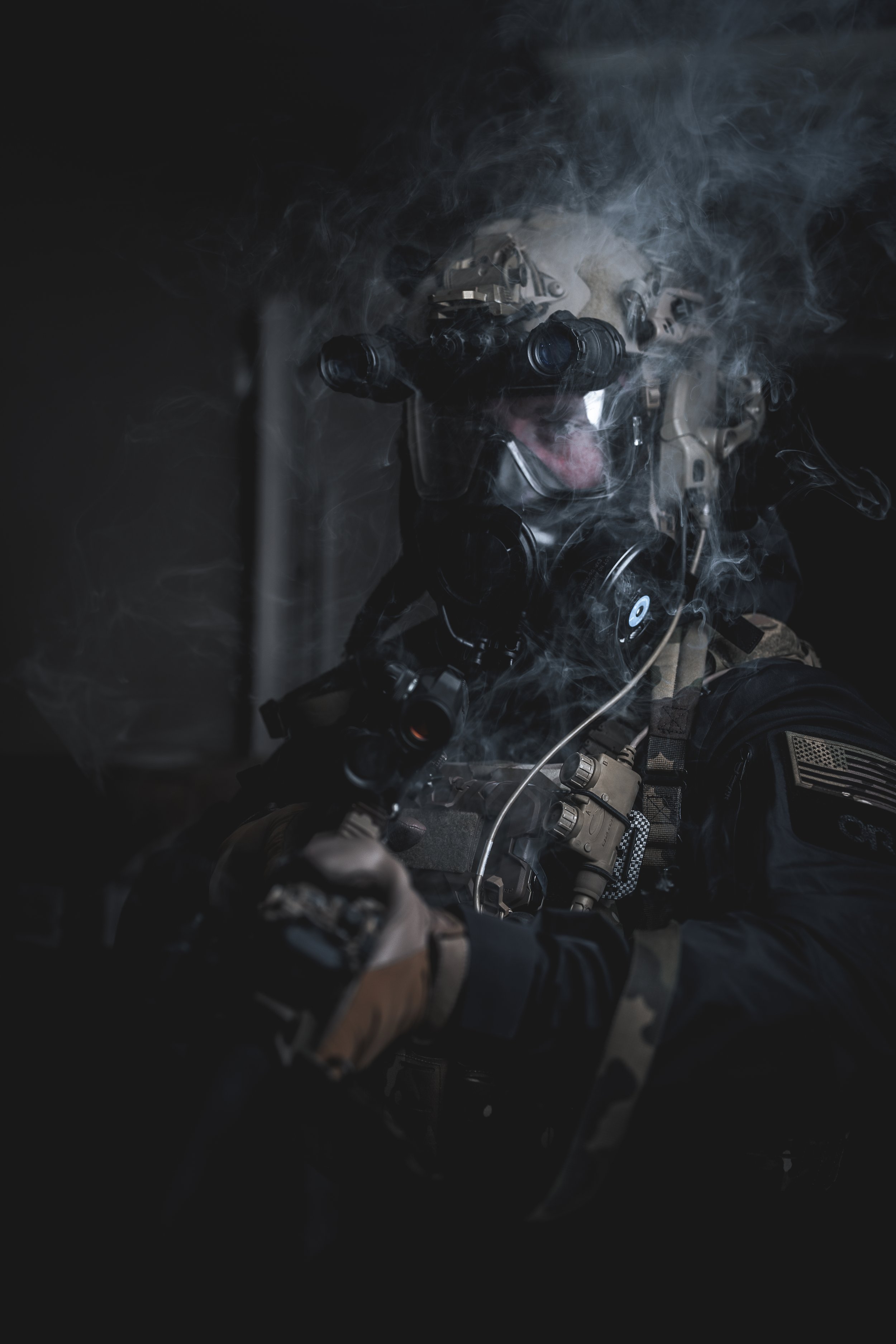 A soldier in tactical gear and night vision goggles surrounded by smoke, holding a weapon.