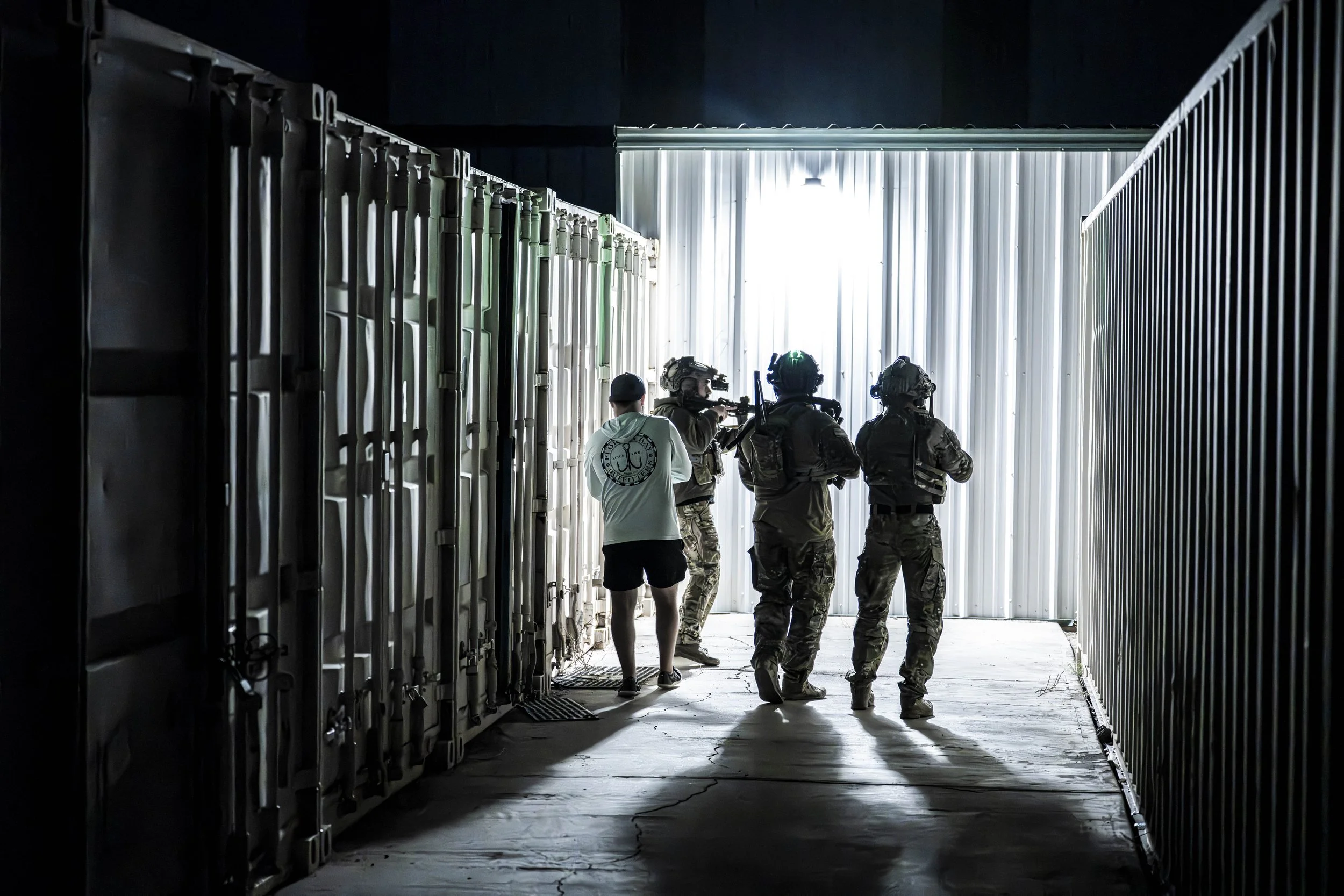 Four uniformed armed personnel and one person in casual clothing walking through a narrow outdoor passage with metal storage units on each side, towards a bright light at the end.