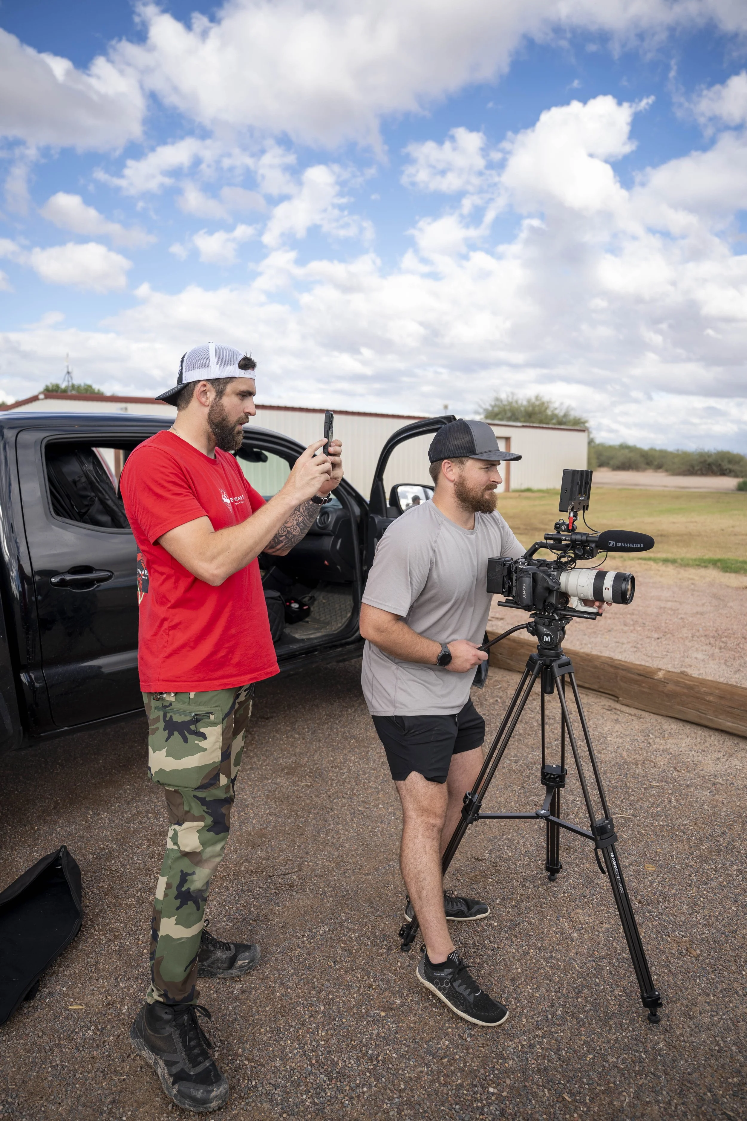 Two men set up a camera on a tripod outdoors near a black pickup truck, with one man taking a photo or recording with a mobile phone, and a rural landscape with a building and partly cloudy sky in the background.