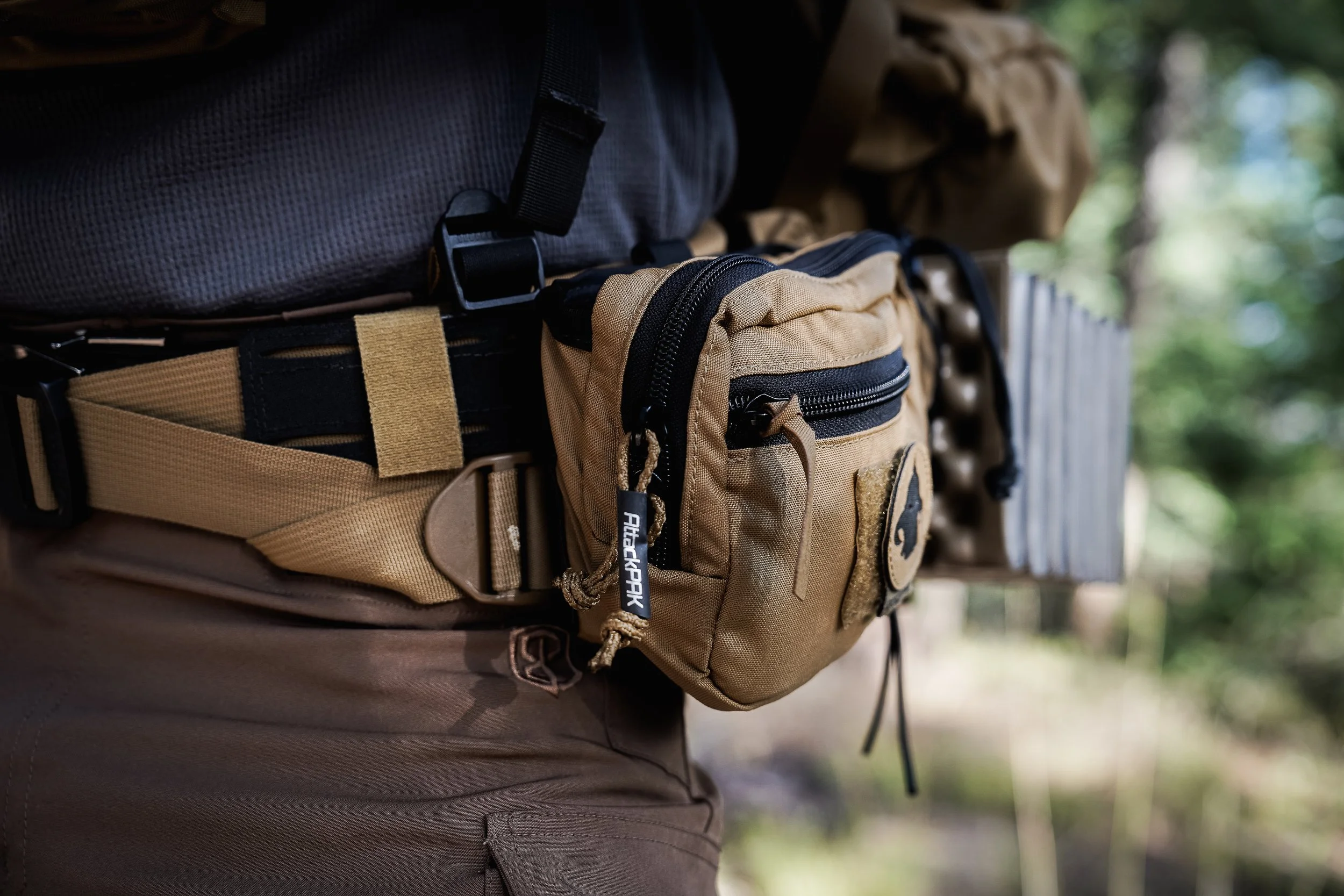 Close-up of a person wearing outdoor gear, including a tan utility belt with pouches and a black buckle, a black backpack, and tan pants, outdoors in a wooded area.
