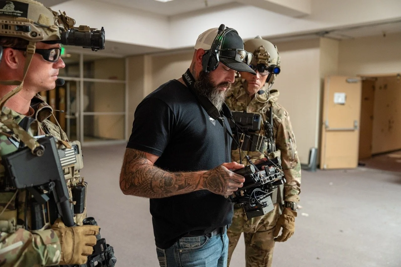Three military personnel in tactical gear and one civilian man with tattoos and headphones are gathered indoors, examining electronic equipment, possibly a drone controller.